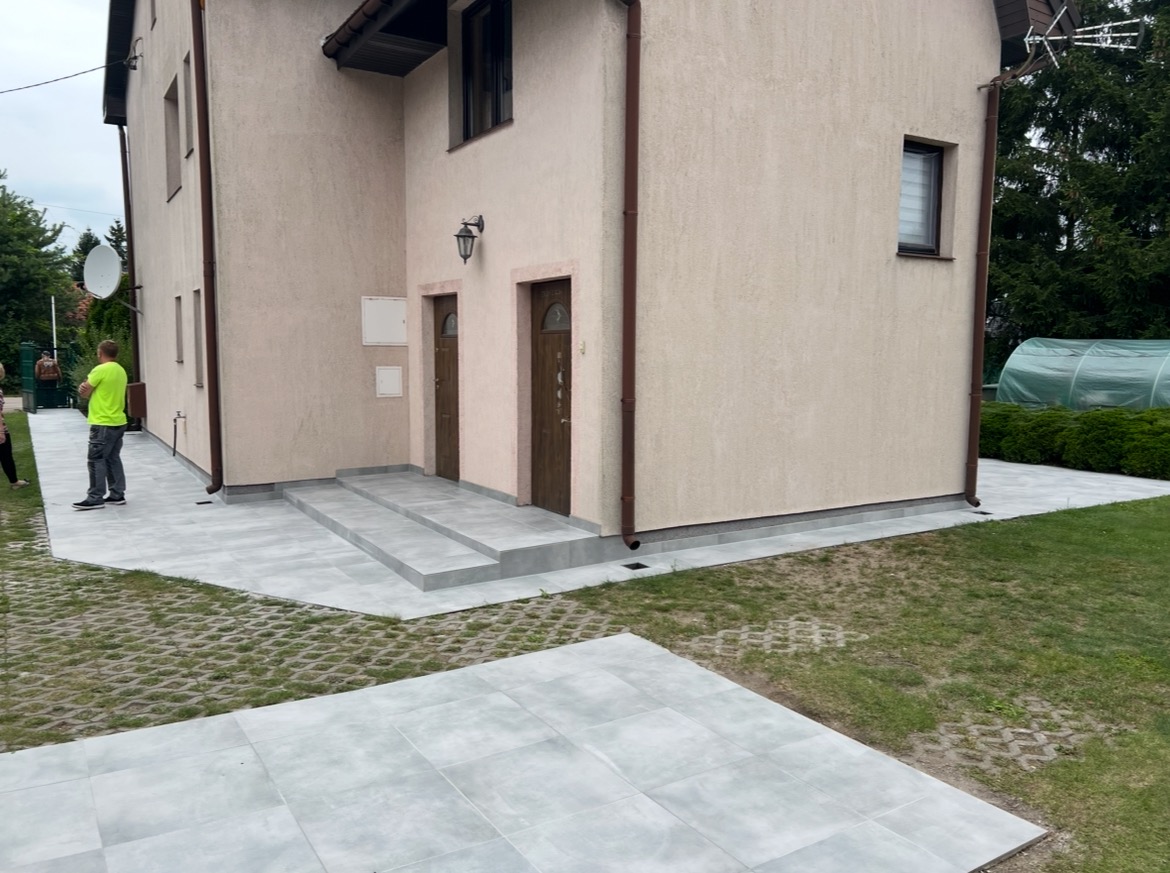 Jasnoszare płytki chodnikowe ułożone przed budynkiem z dwoma wejściami. Widoczne schody z płytek i trawnik z ażurowymi płytami. Oświetlenie dzienne.