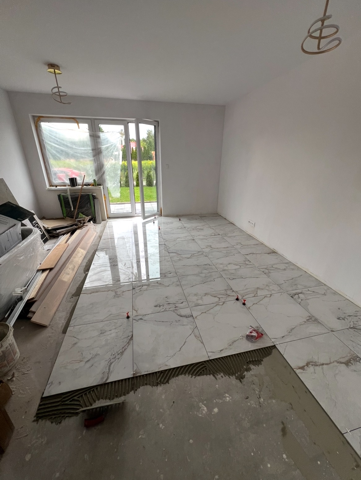 Układanie płytek marmuropodobnych na podłodze w jasnym pokoju. Widoczne narzędzia glazurnicze i krzyżyki dystansowe. Częściowo ułożona podłoga z połyskiem.