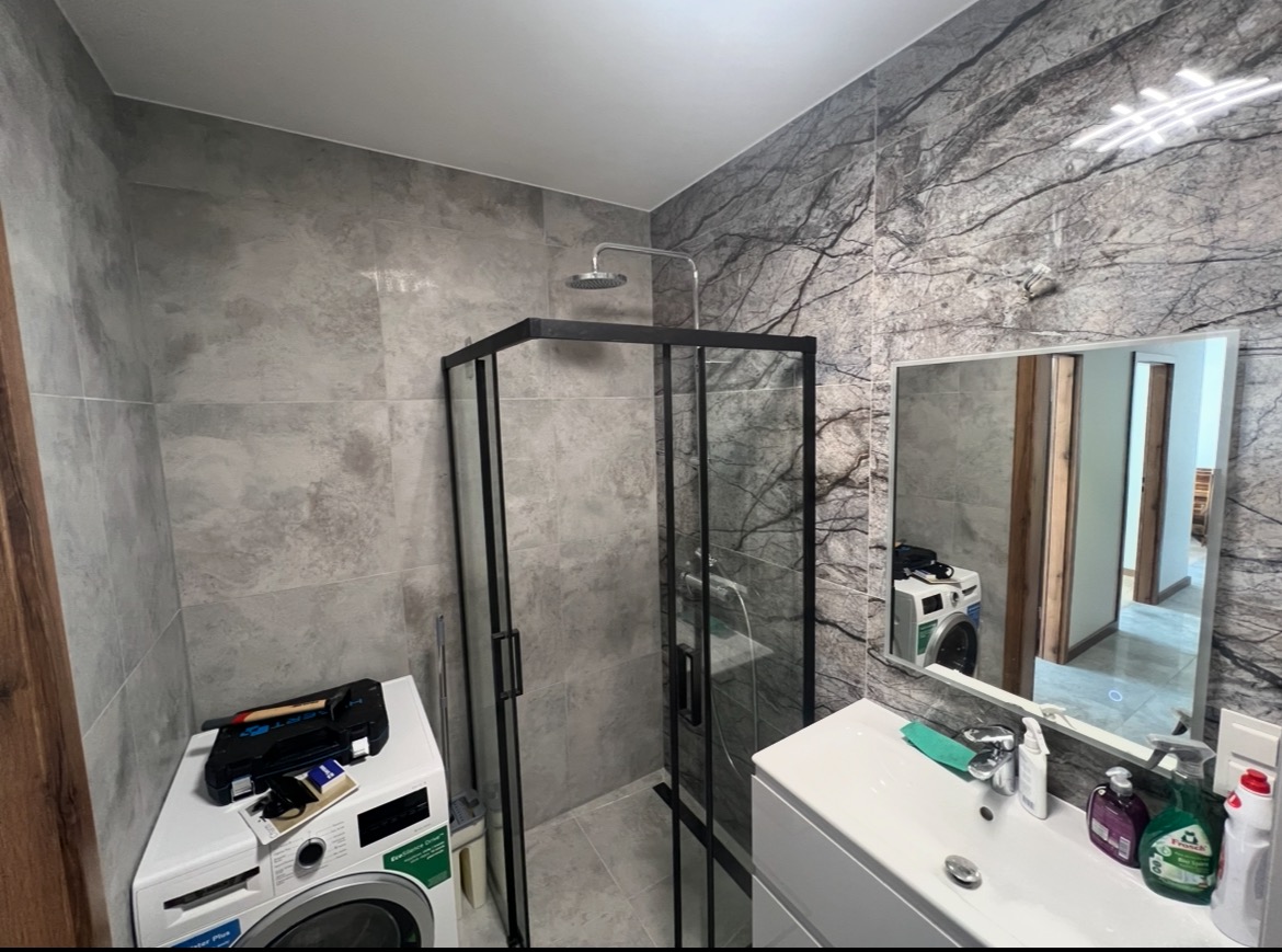 Wykończona łazienka z kabiną prysznicową typu walk-in, umywalką i pralką z narzędziami. Ściany wyłożone płytkami imitującymi marmur.