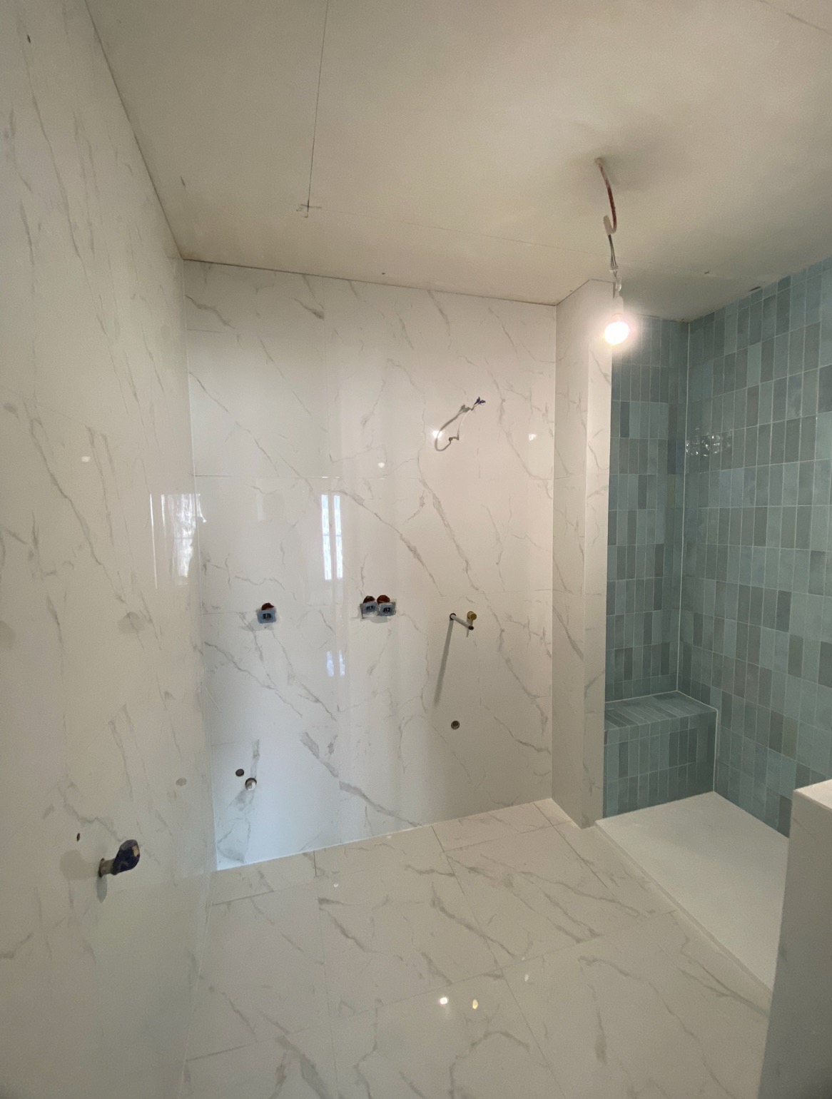 Wnętrze łazienki w trakcie wykańczania: białe płytki imitujące marmur na ścianach i podłodze, fragment kabiny prysznicowej z niebieską mozaiką. Widoczne punkty instalacji.