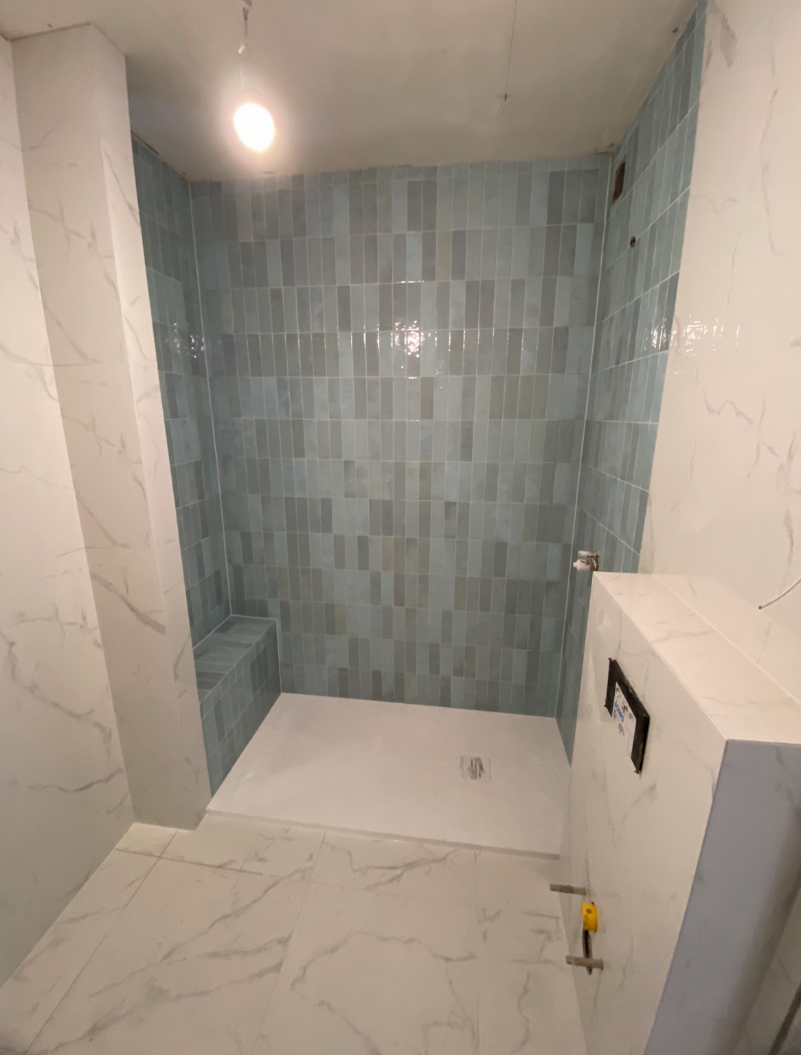 Wykończona kabina prysznicowa z jasnymi, podłużnymi płytkami w odcieniach szarości i błękitu. Widoczny fragment toalety podtynkowej i marmurowe płytki podłogowe.