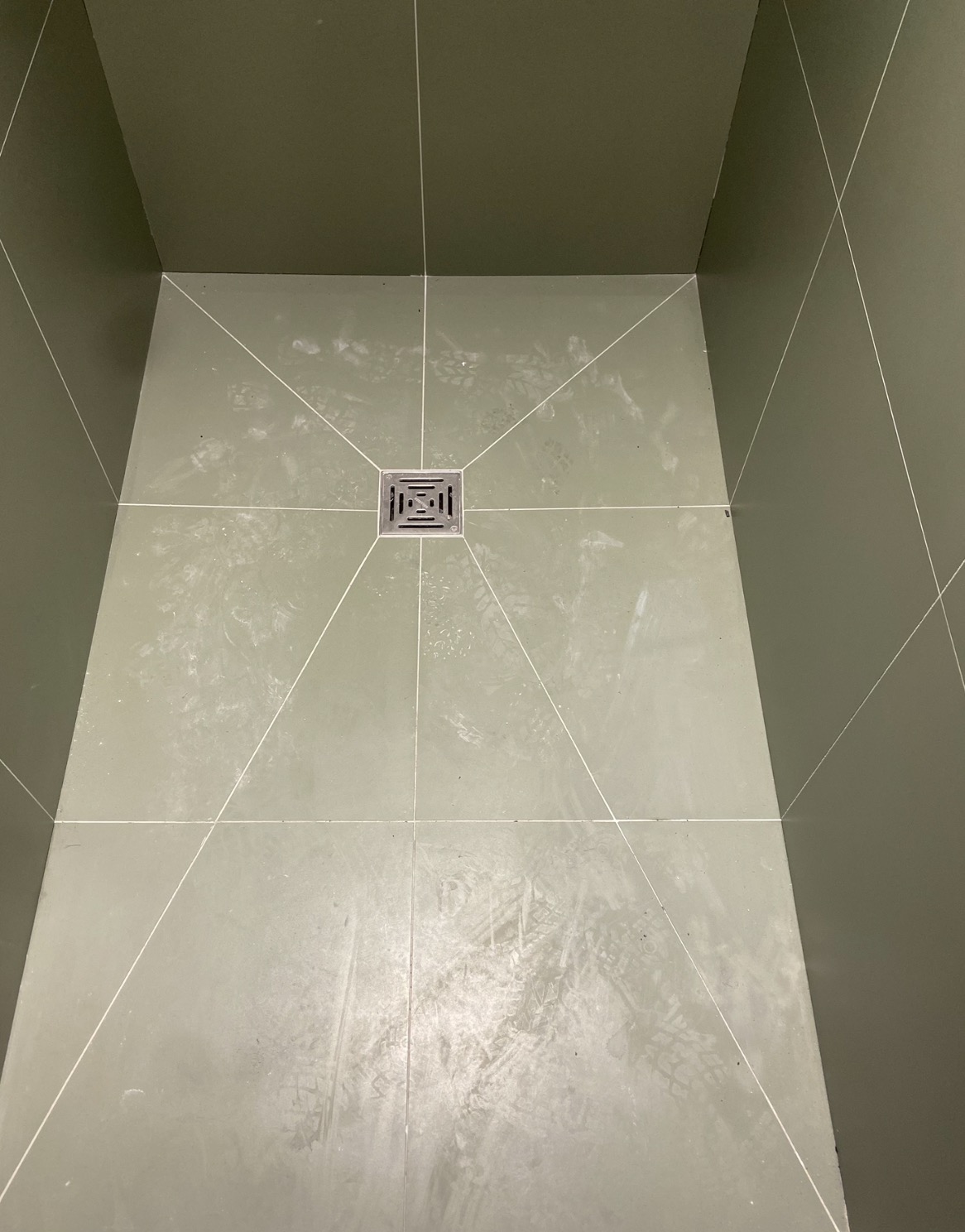 Wykończona kabina prysznicowa z odpływem liniowym, wyłożona szarymi płytkami z białymi fugami. Na podłodze widoczne ślady po pracach wykończeniowych.