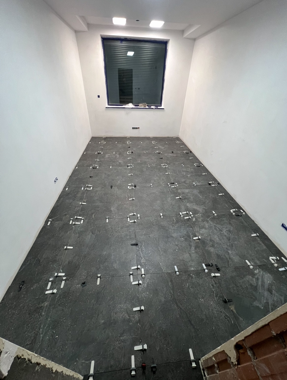 Układanie ciemnych płytek podłogowych w pomieszczeniu z oknem, widoczne krzyżyki dystansowe i kliny poziomujące. Prace wykończeniowe w trakcie realizacji.