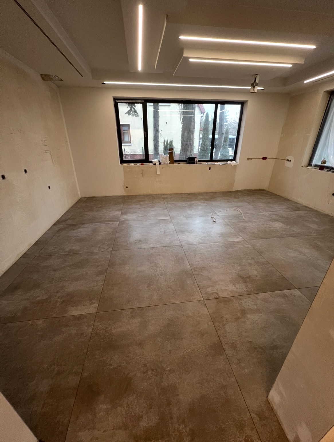Wykończone podłogi z płytek w przestronnym pokoju z oknami. Na ścianach widoczne ślady po instalacjach. Nowoczesne oświetlenie sufitowe.