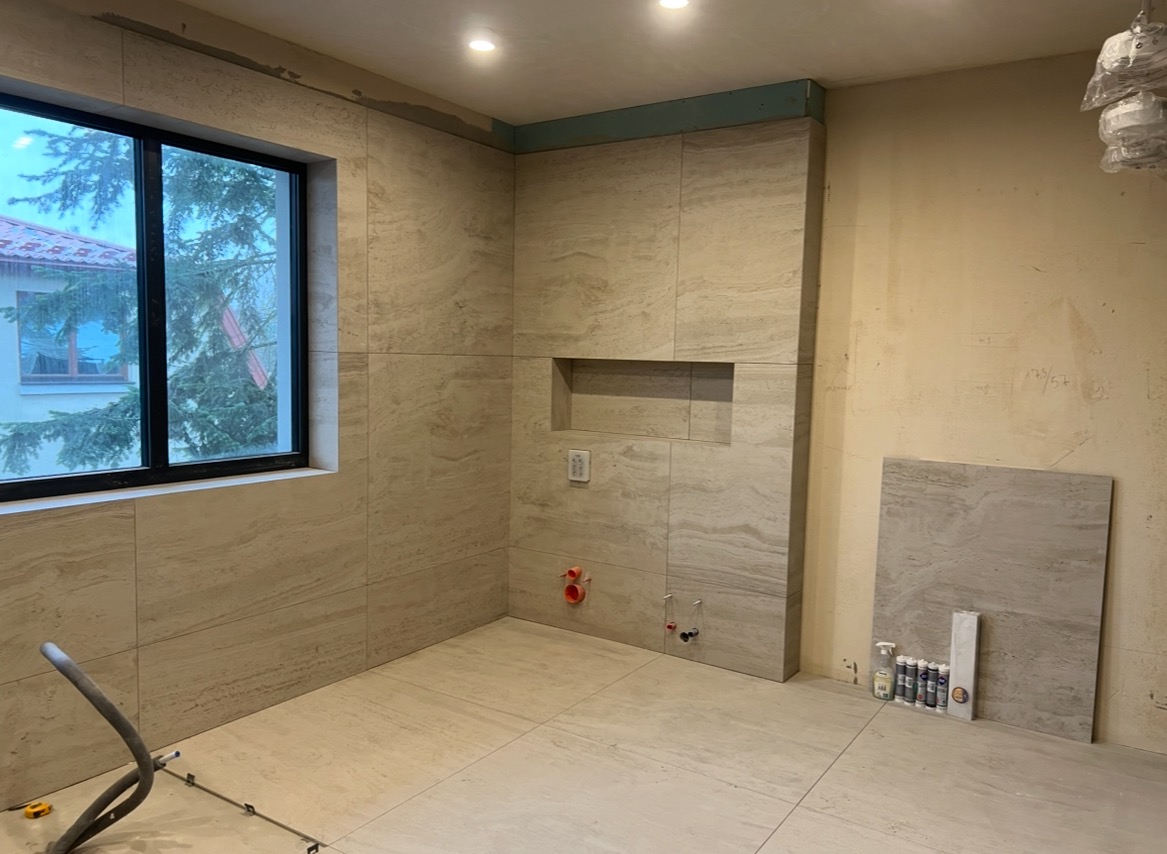 Łazienka w trakcie wykończenia: beżowe płytki imitujące kamień na ścianach i podłodze, okno z widokiem na zewnątrz, widoczne narzędzia i materiały budowlane.