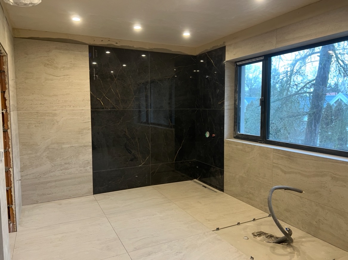 Wykończona łazienka z czarnymi i beżowymi płytkami, widok na okno. Częściowo położona podłoga z wystającą rurą. Nowoczesny design,  oświetlenie punktowe.