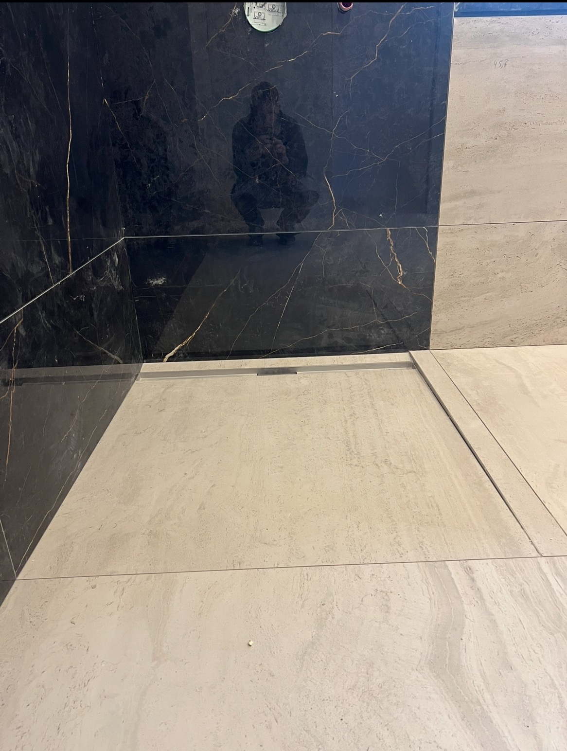 Wykończona strefa prysznicowa z kontrastującymi płytkami: czarny marmur z białymi żyłami na ścianie i beżowe płytki na podłodze. Widoczny odpływ liniowy.