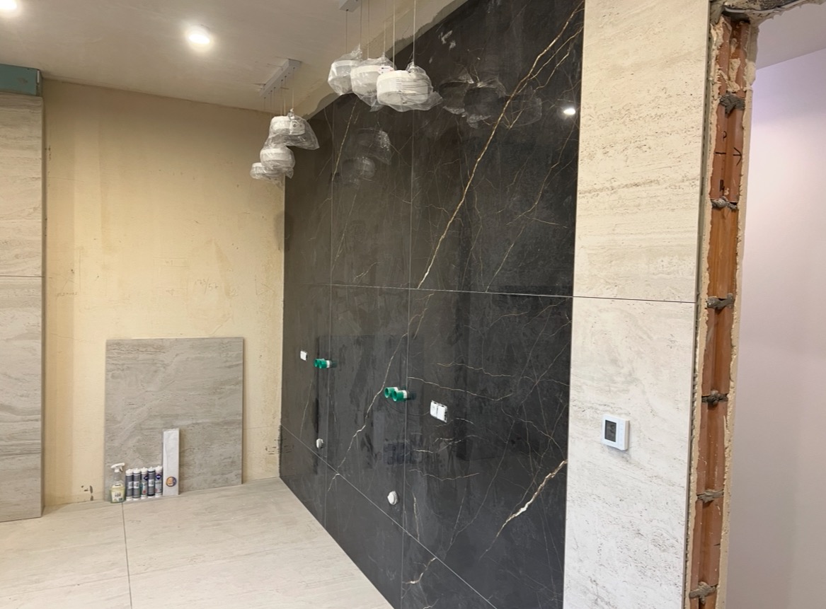 Ściana z płytek imitujących czarny marmur z złotymi żyłami, fragment ściany z jasnego trawertynu, przygotowane instalacje wodne i elektryczne, w tle narzędzia glazurnicze.