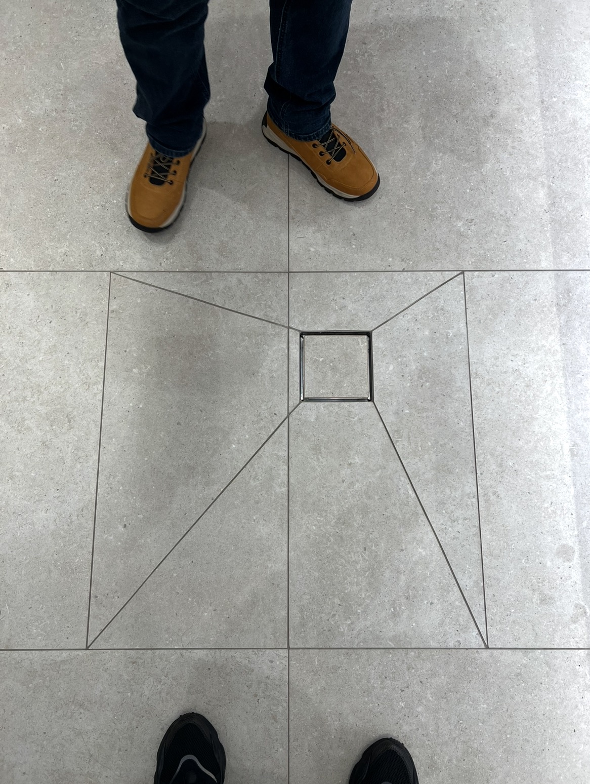 Szare płytki podłogowe z odpływem liniowym w kształcie kwadratu, ułożone z precyzją. Widoczne fragmenty obuwia dwóch osób. Minimalistyczny design.