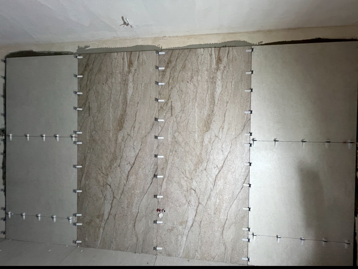Układanie płytek na ścianie z użyciem krzyżyków dystansowych. Częściowo ułożone płytki w odcieniach beżu i szarości, widoczne drobne instalacje elektryczne.