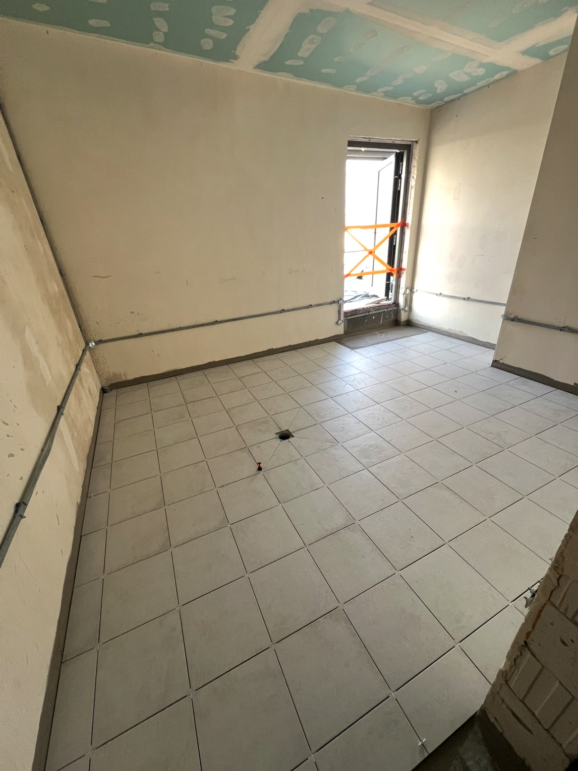 Jasne, kwadratowe płytki podłogowe w pokoju z surowymi ścianami i oknem z pomarańczową taśmą zabezpieczającą. Widoczne akcesoria glazurnicze.