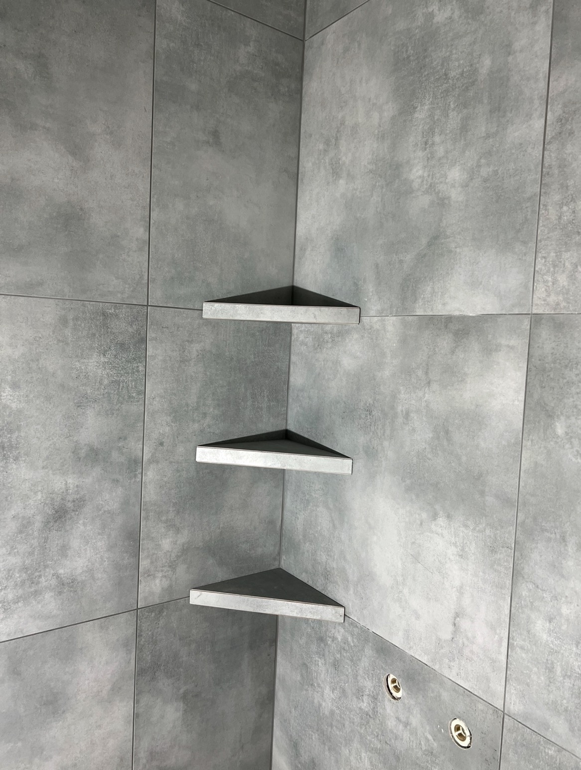 Wykończona ściana kabiny prysznicowej z szarych płytek z trzema półkami narożnymi w kształcie trójkąta, umieszczonymi jedna nad drugą.