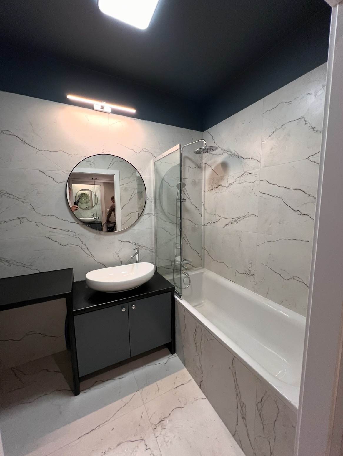 Wykończona łazienka z wanną, szklanym parawanem, umywalką na szafce i lustrem. Płytki imitujące marmur na ścianach i podłodze, ciemny sufit.