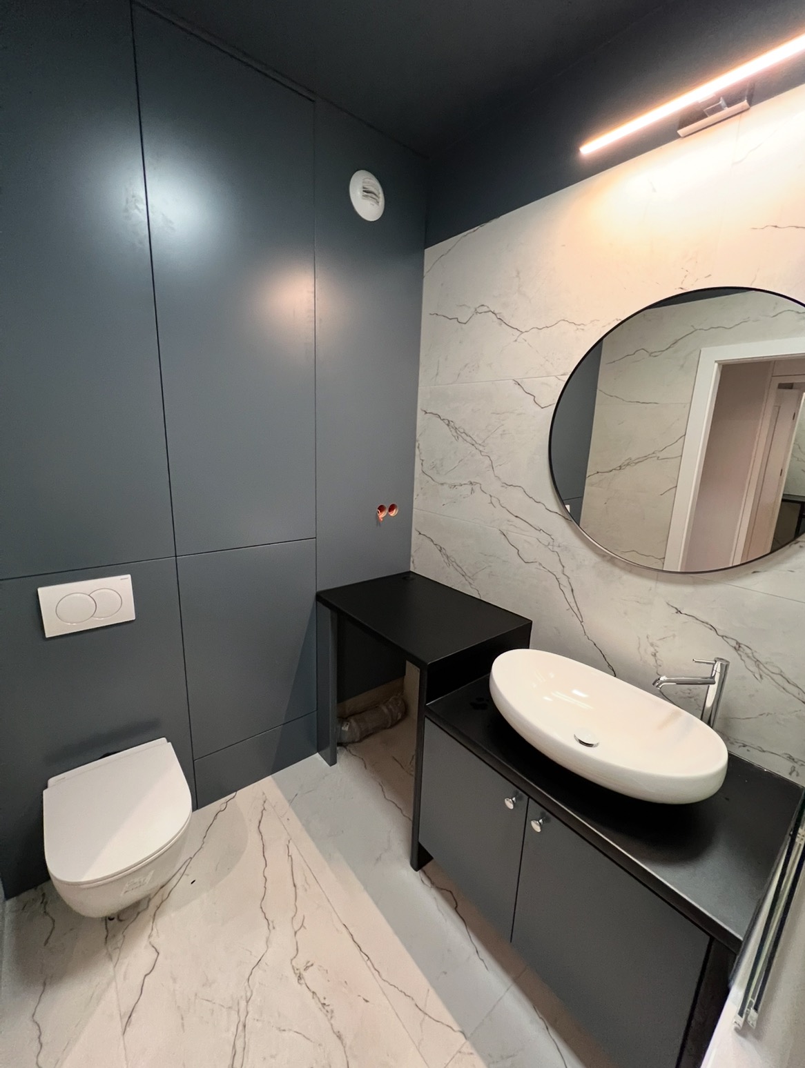 Elegancka łazienka z szarymi płytami, okrągłym lustrem i białą umywalką nablatową. Minimalistyczny design, marmurowe płytki i nowoczesne oświetlenie.