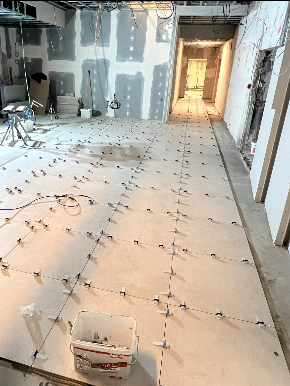 Układanie płytek podłogowych z użyciem klipsów poziomujących w długim pomieszczeniu w trakcie remontu. Widoczny kubeł z klejem i narzędzia.