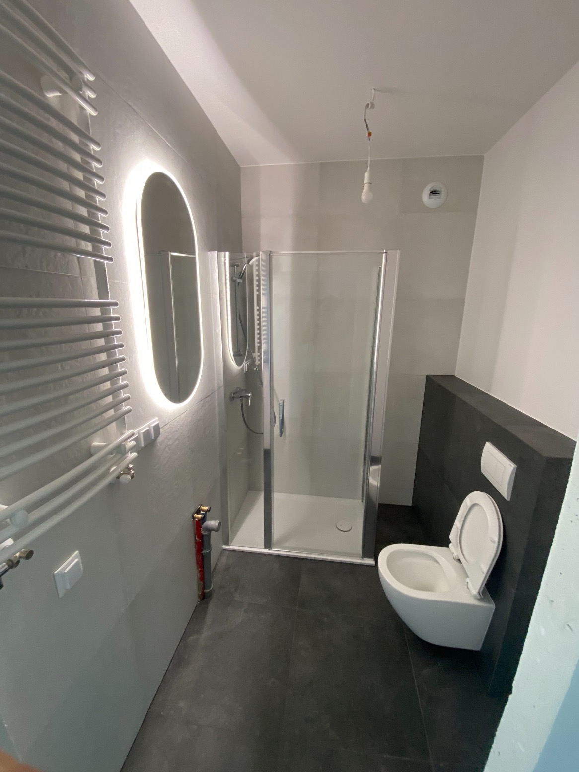 Nowoczesna łazienka po remoncie: kabina prysznicowa, podświetlane lustro, grzejnik drabinkowy, toaleta podwieszana i ciemna podłoga. Widok z góry.