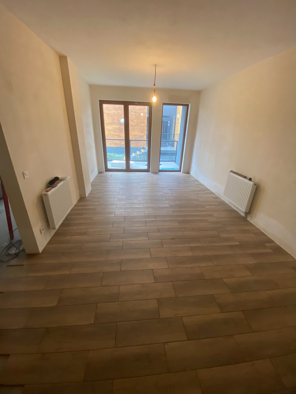 Ukończone układanie podłogi z płytek imitujących drewno w jasnym pomieszczeniu z oknami balkonowymi i grzejnikami. Widoczne niedoskonałości wykończenia ścian.