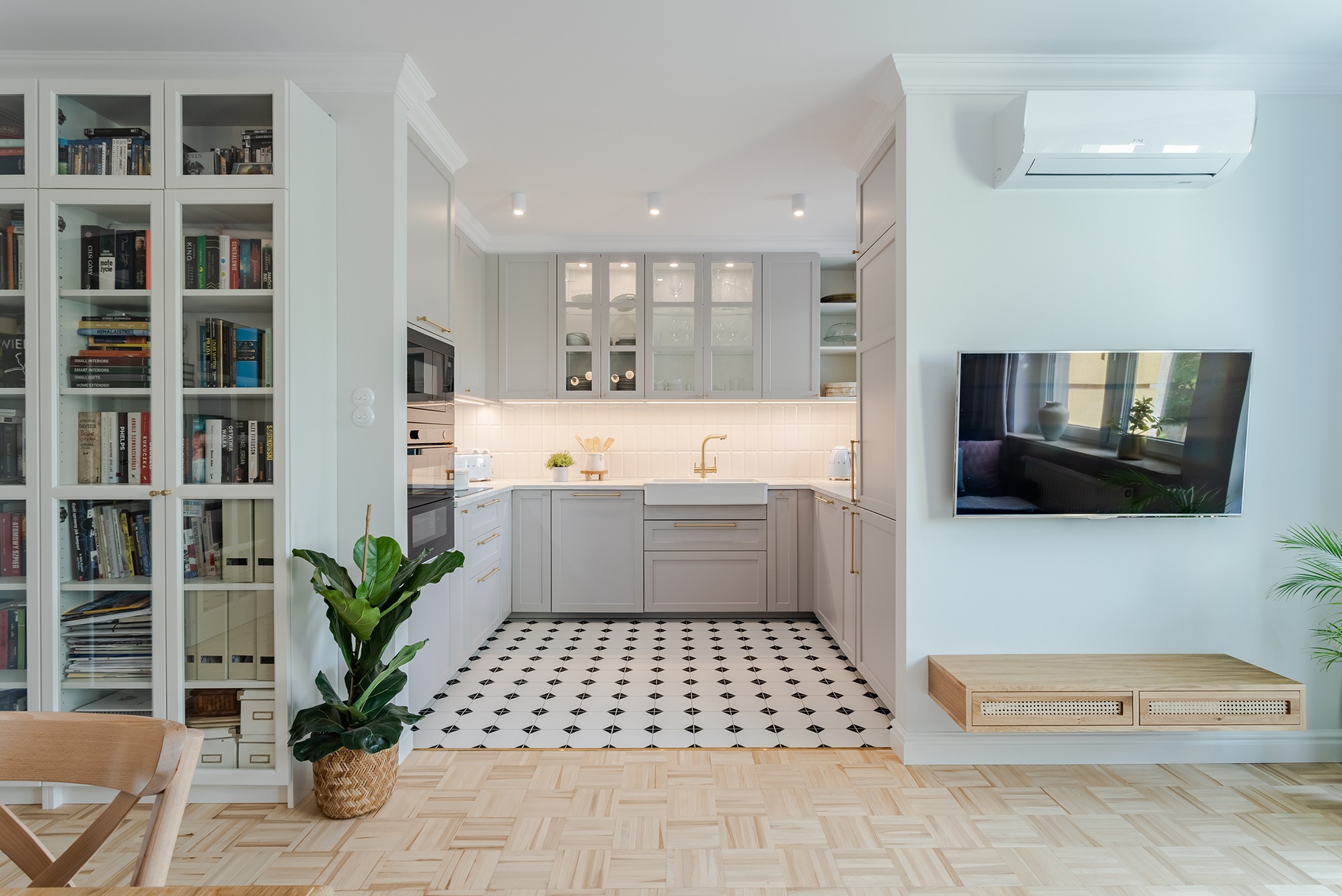 Jasna kuchnia w stylu retro z szarymi szafkami, złotymi uchwytami i armaturą, białą ceramiką i czarno-białą podłogą w geometryczny wzór, widok z salonu z parkietem, regałem z książkami...