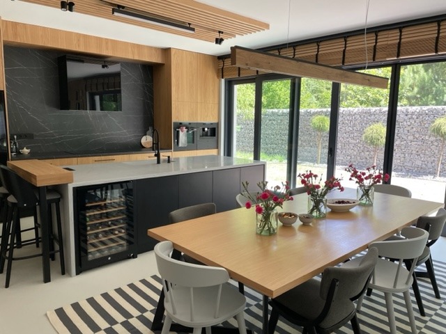 Nowoczesna kuchnia z wyspą i barkiem, połączona z jadalnią, z dużym oknem na ogród z kamiennym murem i kulistymi drzewami.