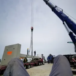 Widok z perspektywy leżącego człowieka na montaż betonowego elementu konstrukcyjnego za pomocą dźwigu, dwóch pracowników w kaskach nadzoruje proces.