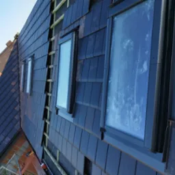 Montaż ciemnej dachówki ceramicznej z oknami dachowymi, widoczne drewniane łaty konstrukcyjne i częściowo ułożony dach.