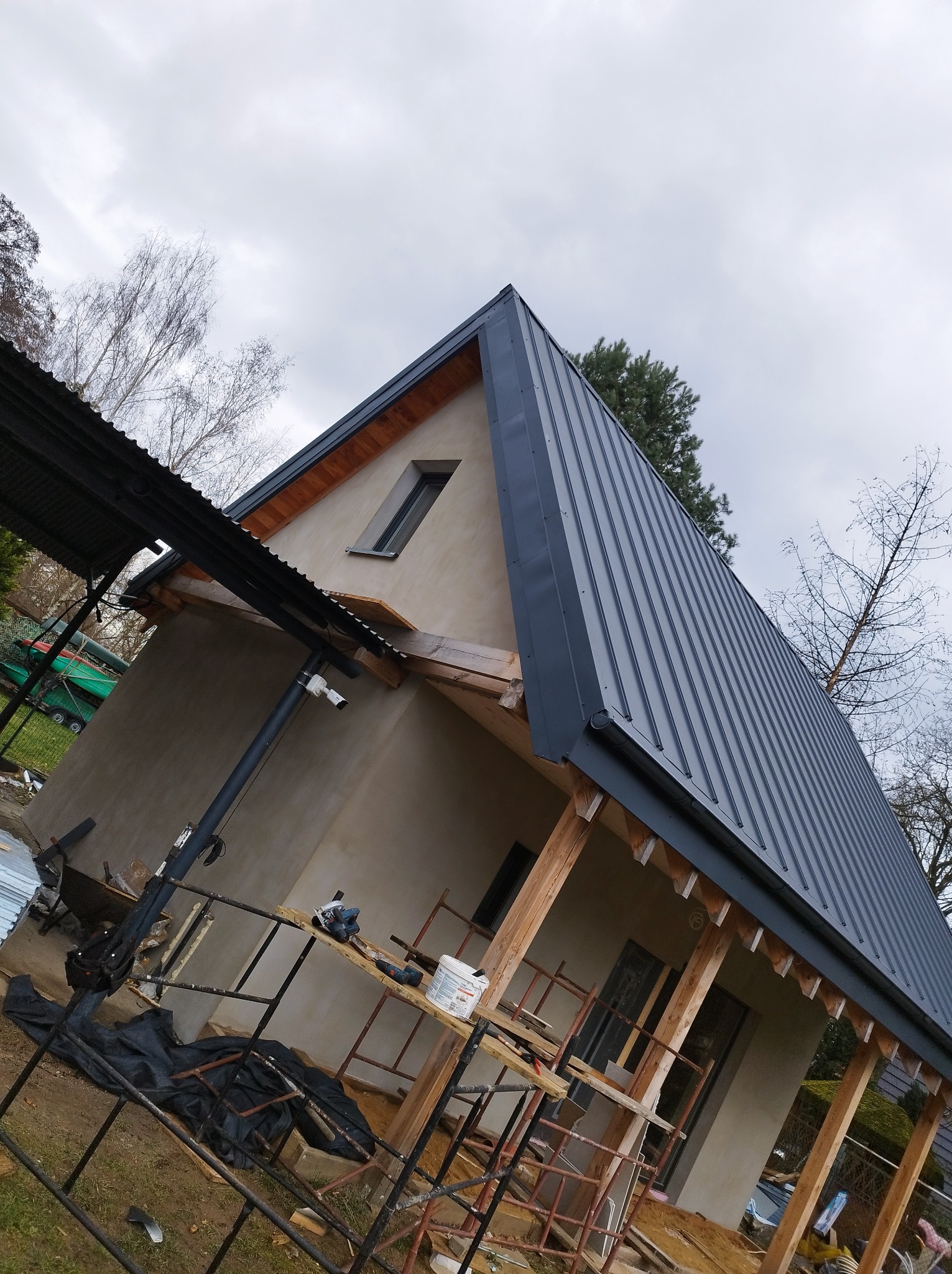 Dom w trakcie budowy z nowym, grafitowym dachem blaszanym, widoczne rusztowania i narzędzia.