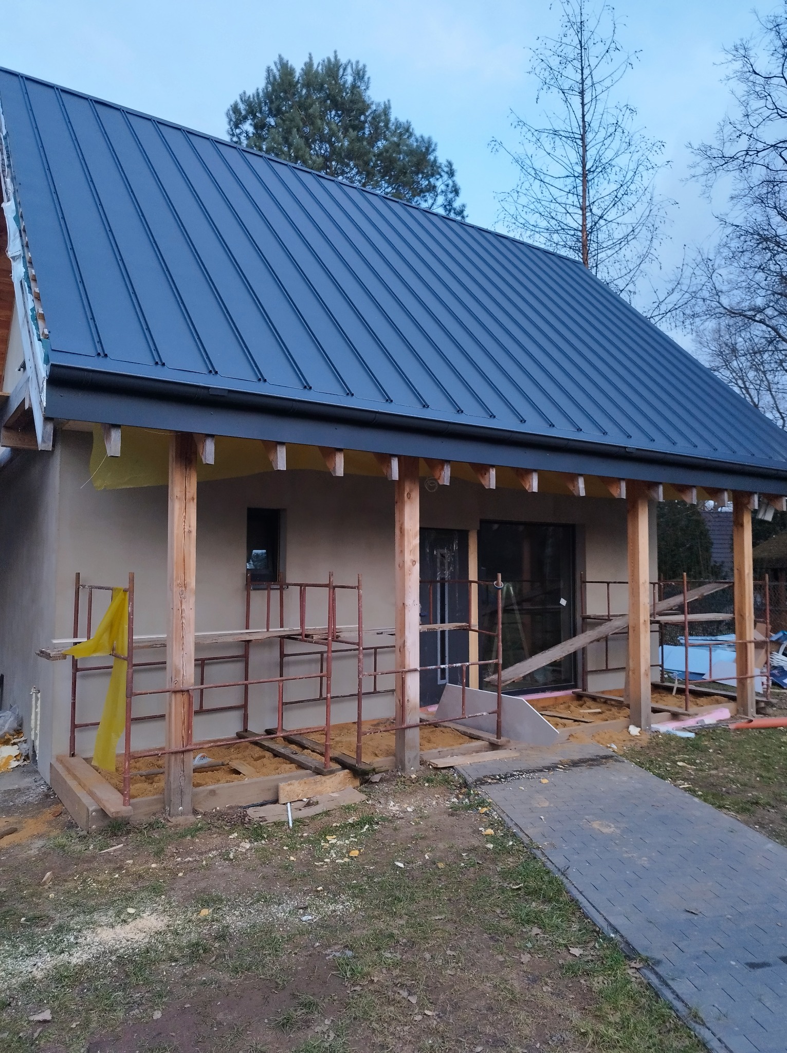 Dom w trakcie budowy z nowym, grafitowym dachem z blachy na rąbek stojący, widoczne rusztowania, ocieplenie i elementy konstrukcyjne werandy.