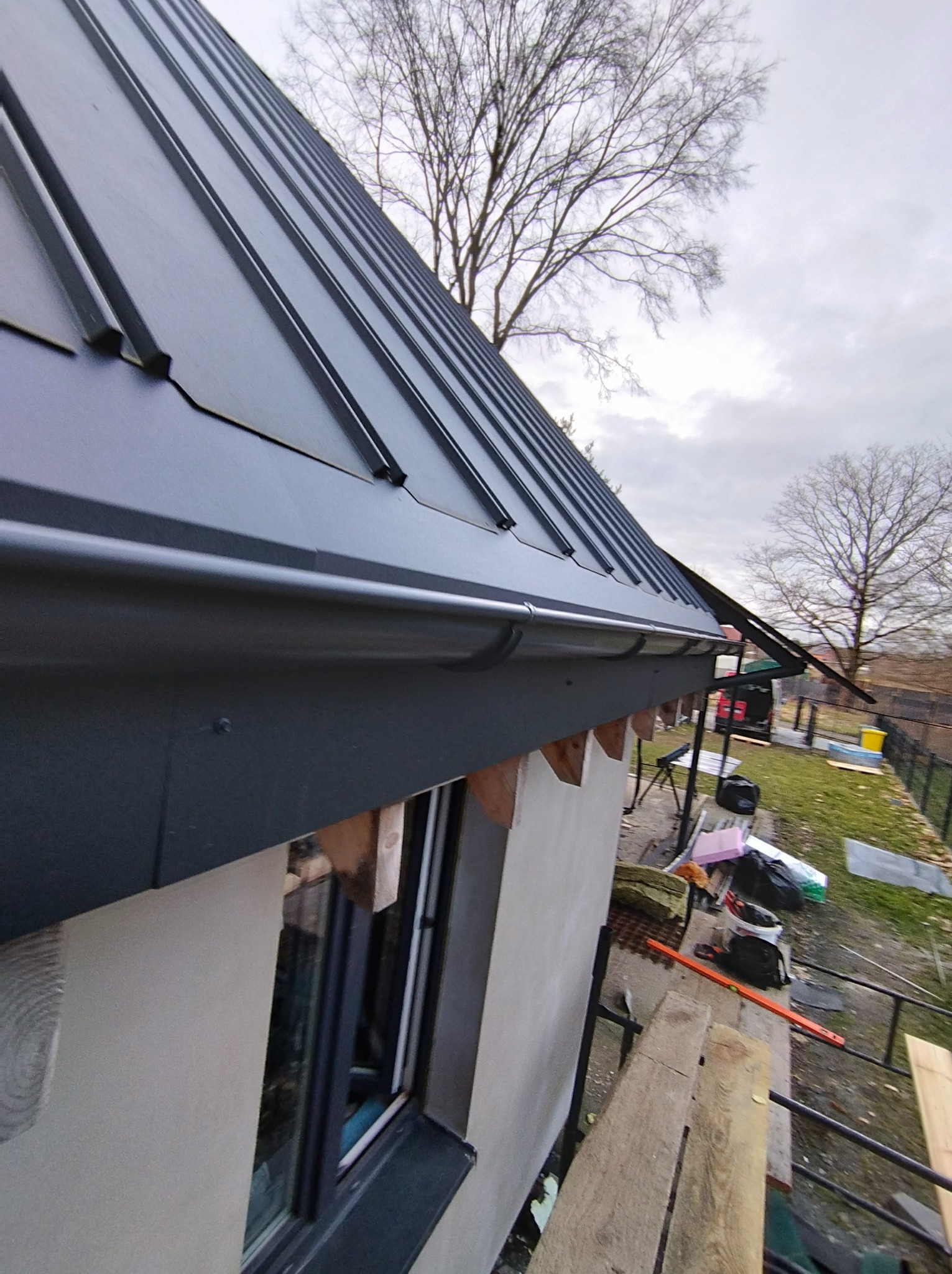 Widok z dołu na nowo zamontowany, ciemny, blaszany dach z rynnami, drewniane elementy konstrukcji, rusztowanie i narzędzia budowlane w tle.