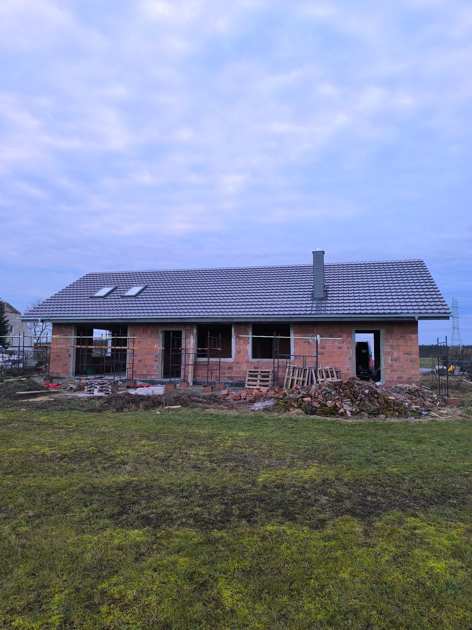 Surowy stan domu jednorodzinnego z czerwonej cegły, z szarą dachówką i dwoma oknami dachowymi, w trakcie budowy, na tle pochmurnego nieba i zielonej trawy.