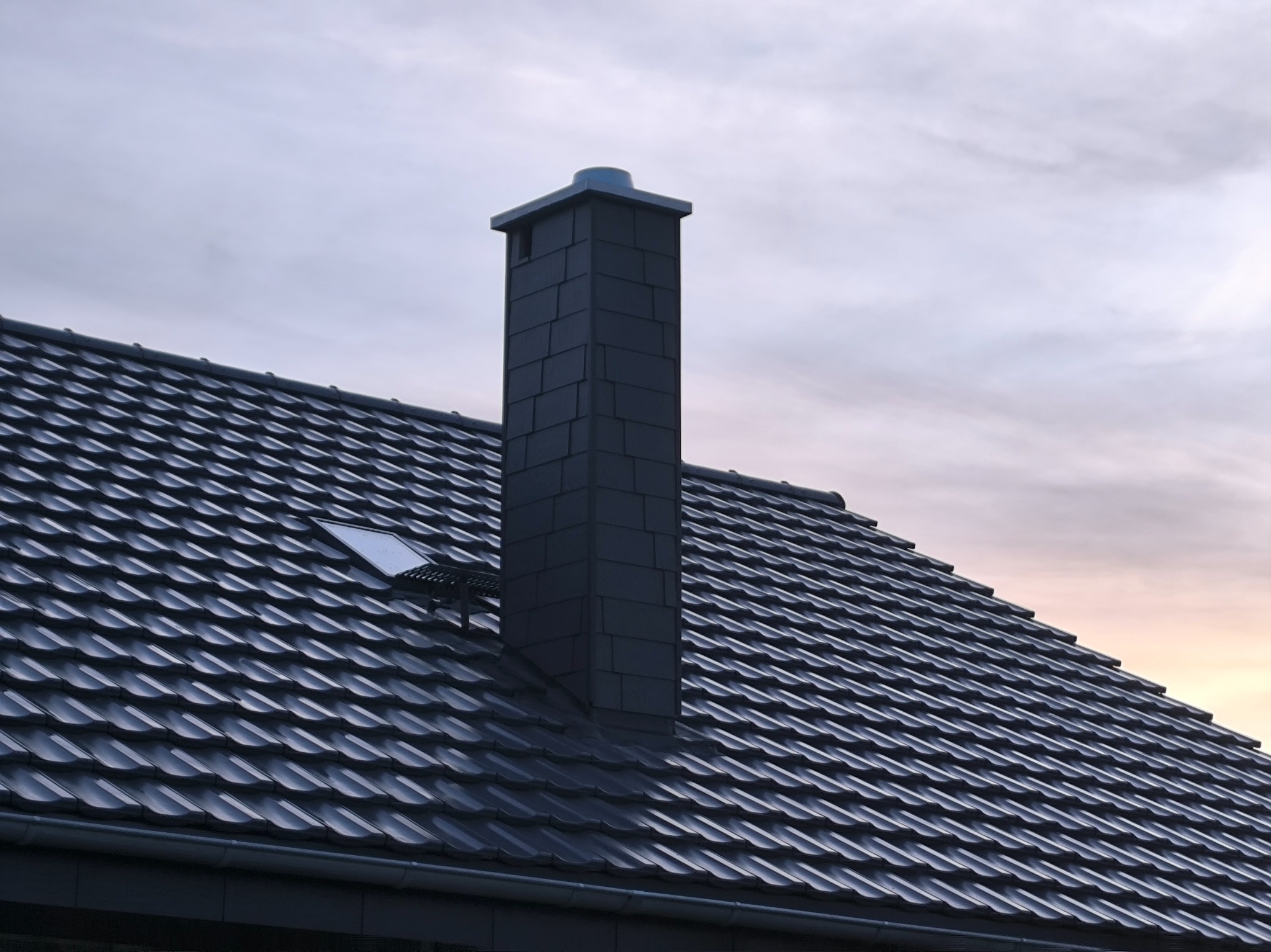 Grafitowy dach z dachówki ceramicznej z kominem w tym samym kolorze, widoczny fragment okna dachowego. Tło stanowi pochmurne niebo o zmierzchu.