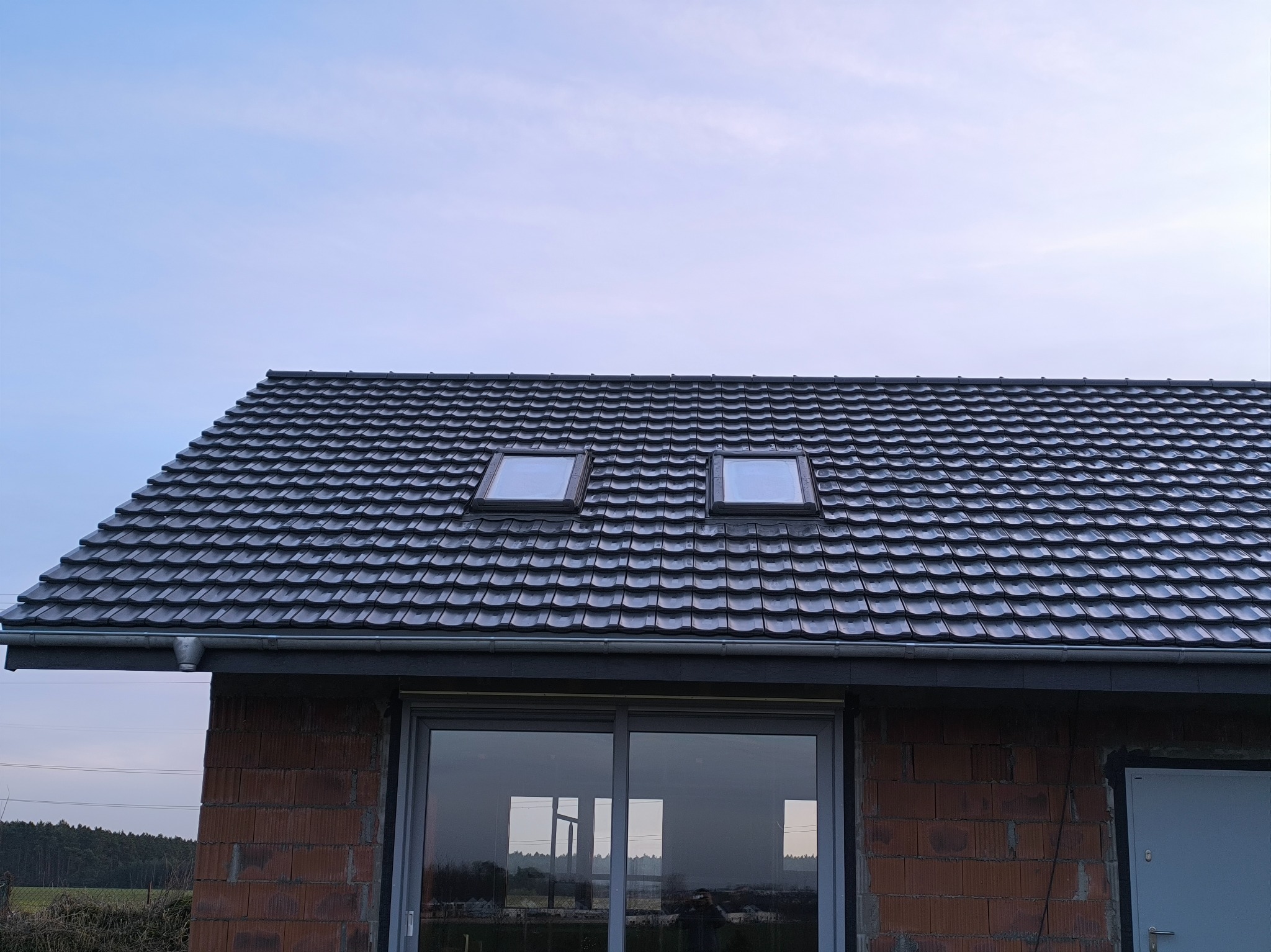 Ciemna dachówka ceramiczna z dwoma oknami dachowymi na nowym domu w stanie surowym otwartym, widoczne rynny i okno.