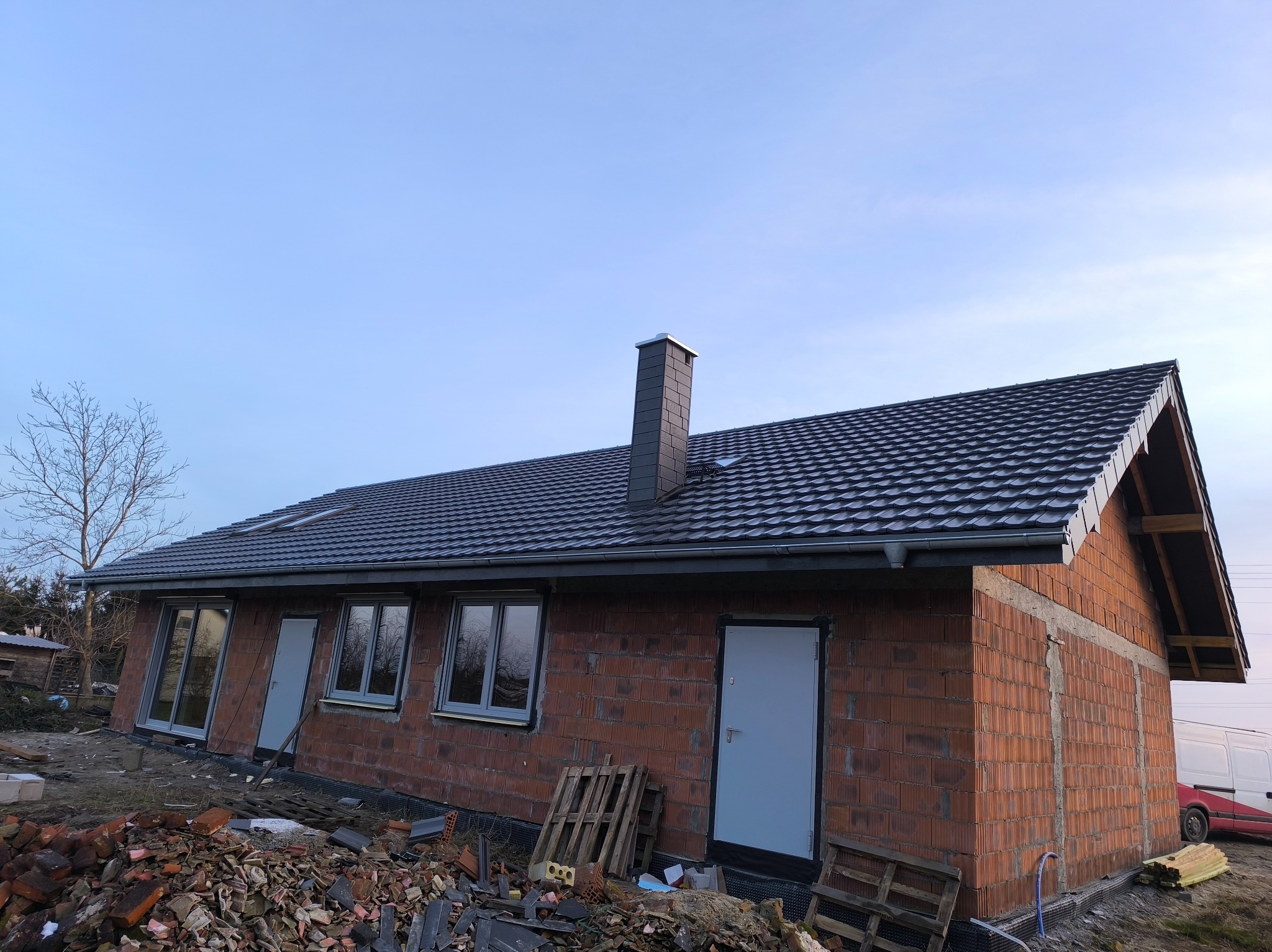Dom w trakcie budowy z ciemnym dachem i kominem. Widoczne cegły, okna i drzwi. Na pierwszym planie gruz i palety. Niebo błękitne.