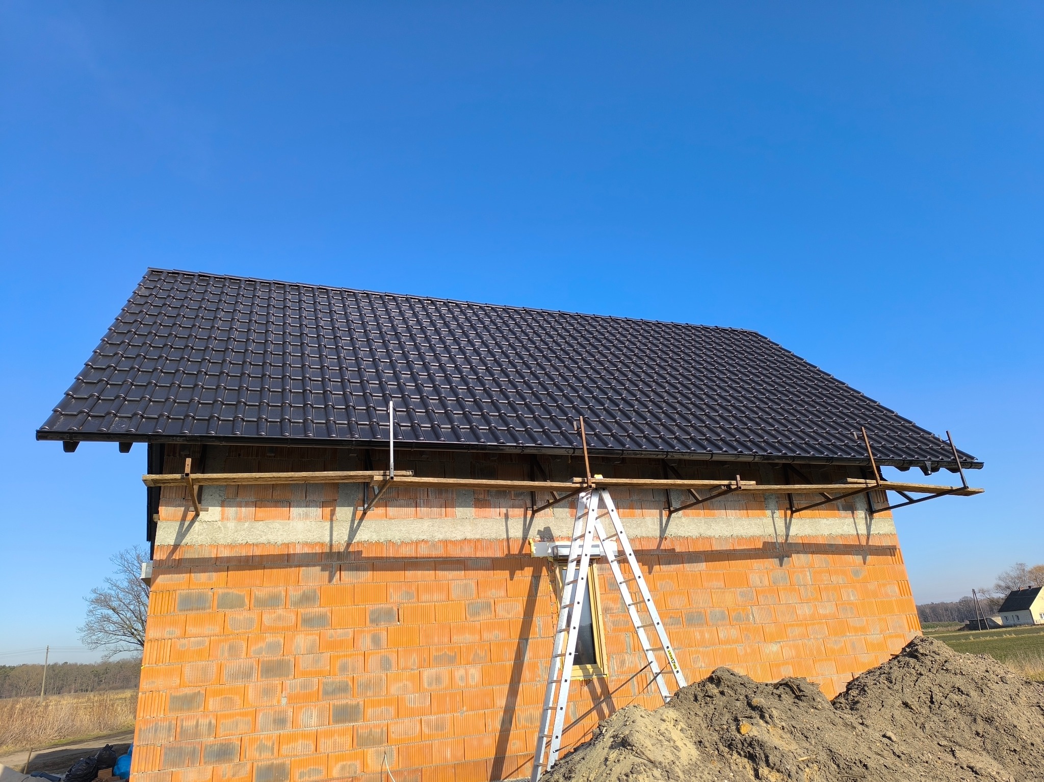 Nowy dach z czarnej dachówki ceramicznej, zamontowany na budynku w Namysłowie. Widoczne rusztowania i drabina, błękitne niebo w tle. Dachówka ułożona równo, z dbałością o detale.