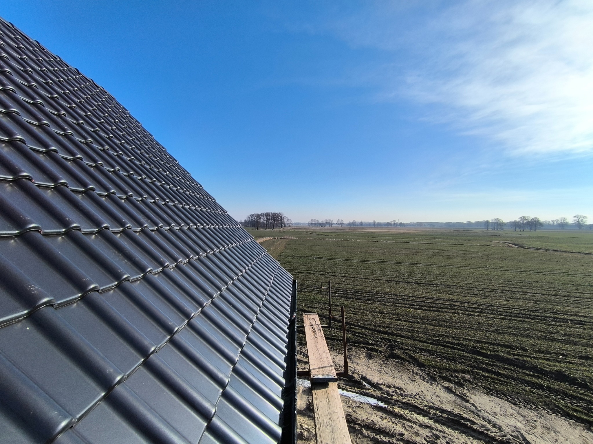 Błyszczący, grafitowy dach z dachówki ceramicznej, widziany z góry, z rusztowaniem i rozległym polem w tle pod błękitnym niebem.