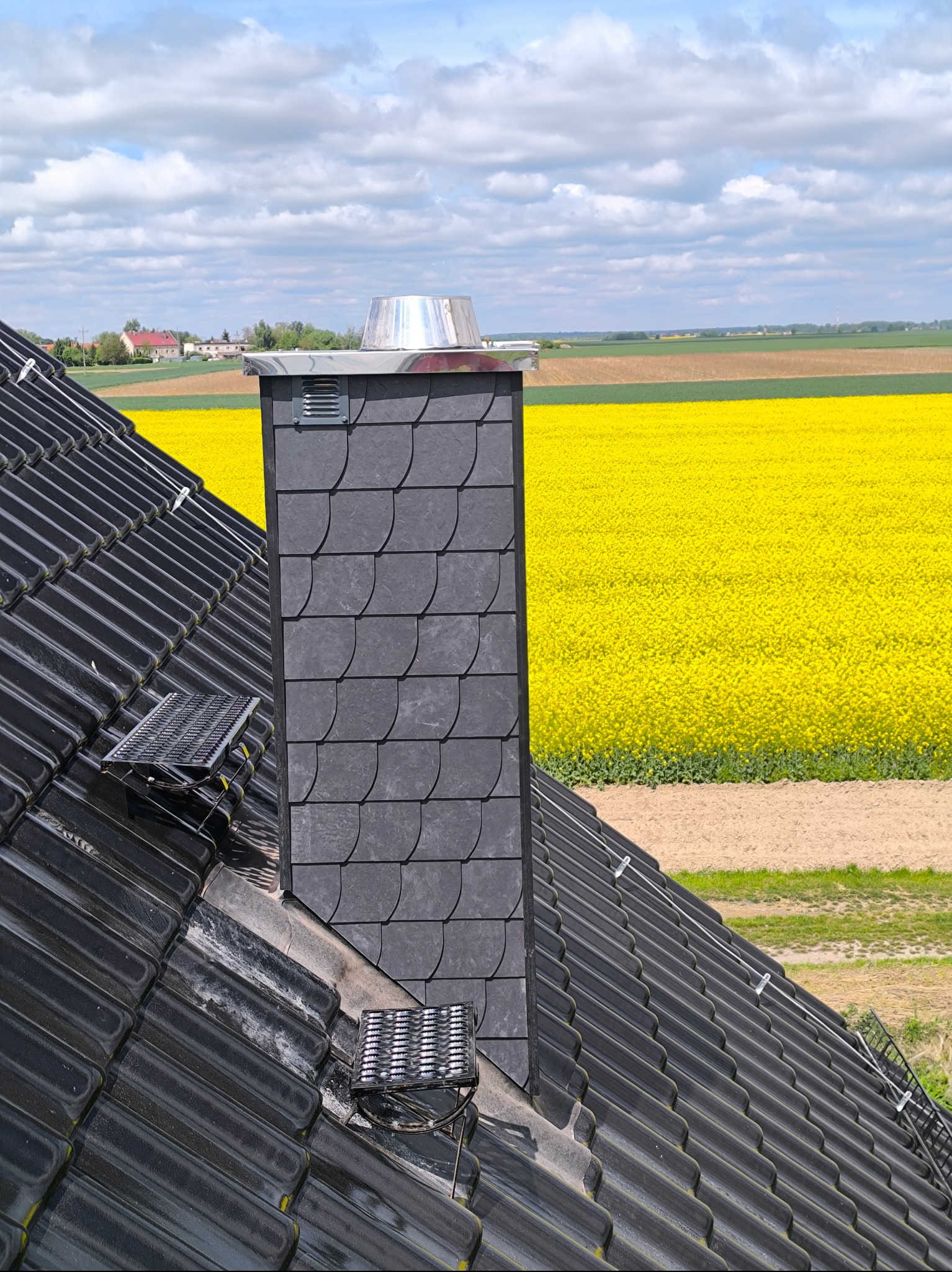 Czarny dach z dachówki ceramicznej, komin z łupka z metalowym zakończeniem na tle kwitnącego pola rzepaku i zabudowań wiejskich w oddali.