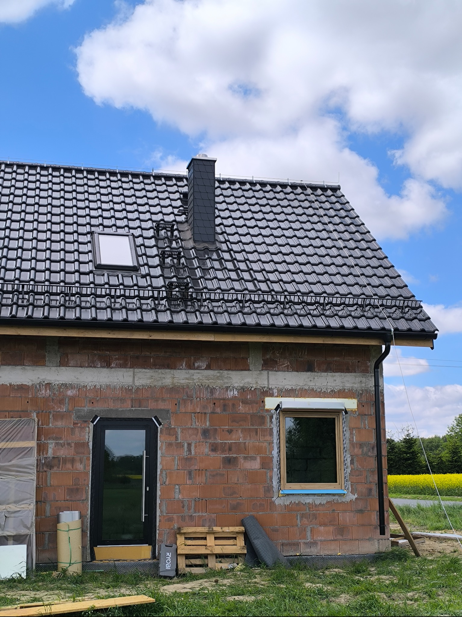 Dom w trakcie budowy z czarnym dachem z dachówki ceramicznej, kominem i oknem dachowym. Widoczne niezadaszone ściany z czerwonej cegły.