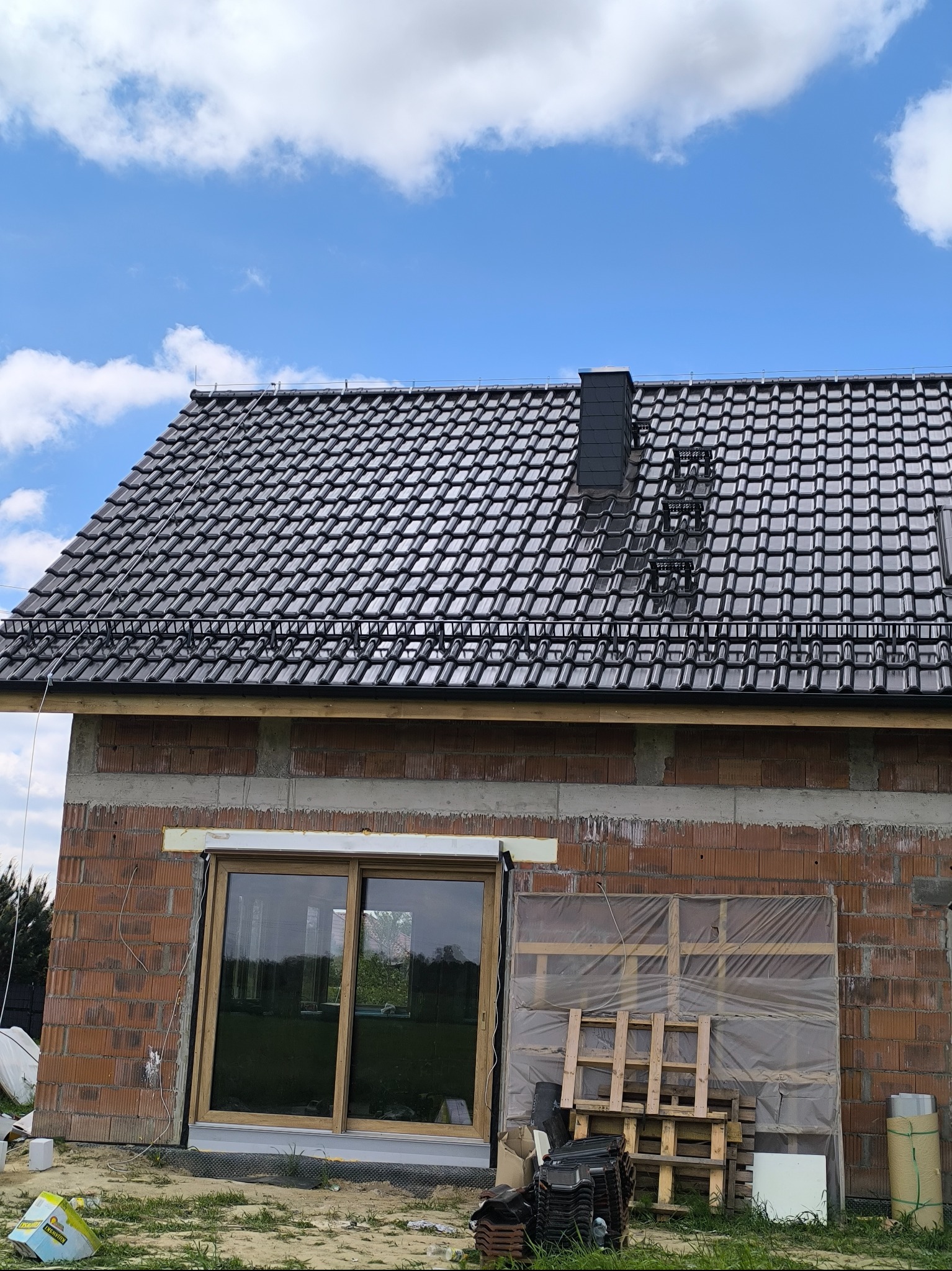 Nowy, błyszczący dach z czarnej dachówki ceramicznej na surowym budynku z czerwonej cegły, widok z dołu, błękitne niebo z chmurami w tle.