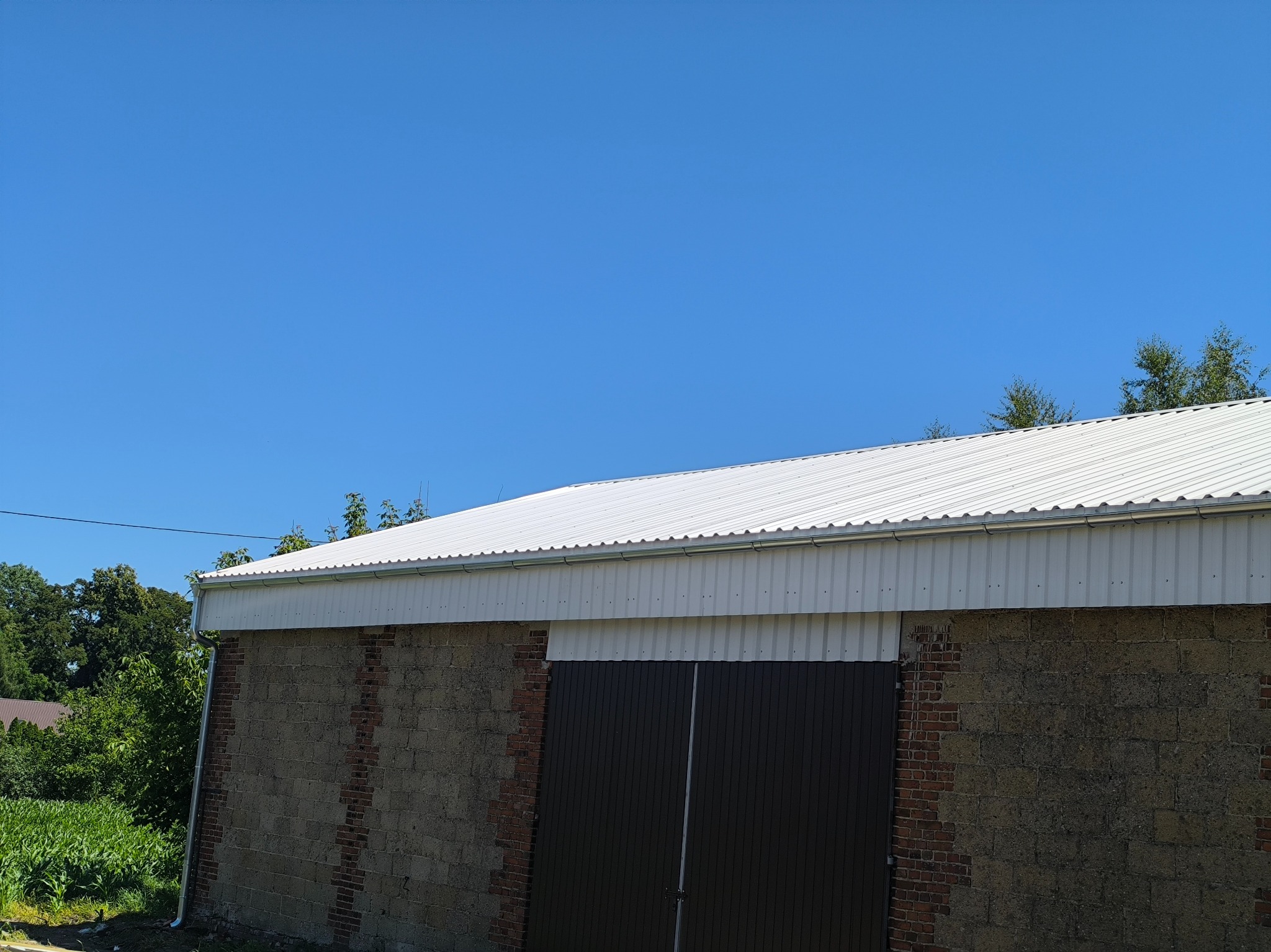 Biały, blaszany dach z rynnami na murowanym budynku gospodarczym pod błękitnym niebem. Widoczne drzewa w tle i fragment bramy wjazdowej.