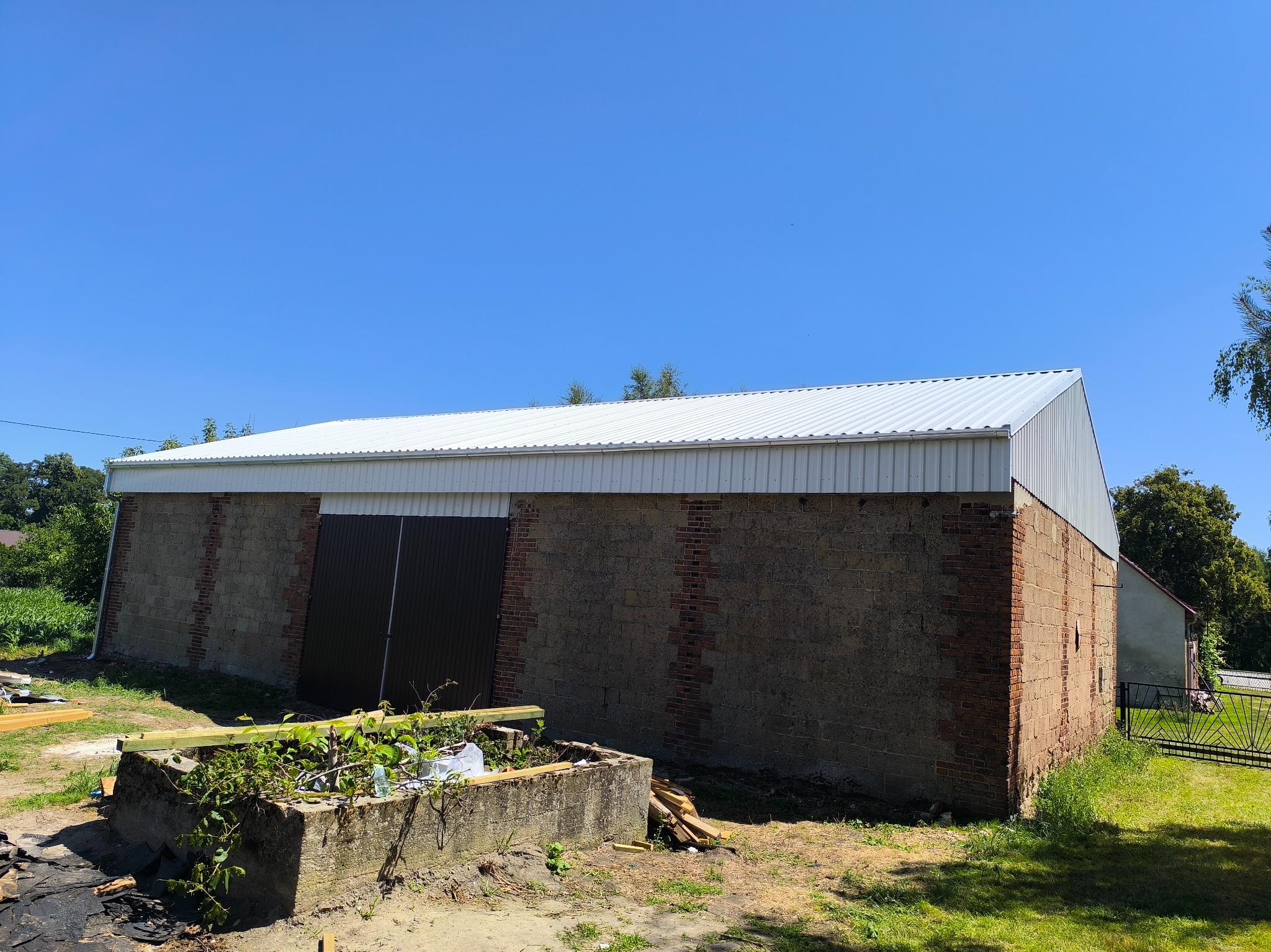 Budynek gospodarczy z nowym, białym dachem z blachy trapezowej, murowane ściany z ceglanymi wstawkami, ciemne wrota. Na pierwszym planie betonowa konstrukcja porośnięta roślinnością.