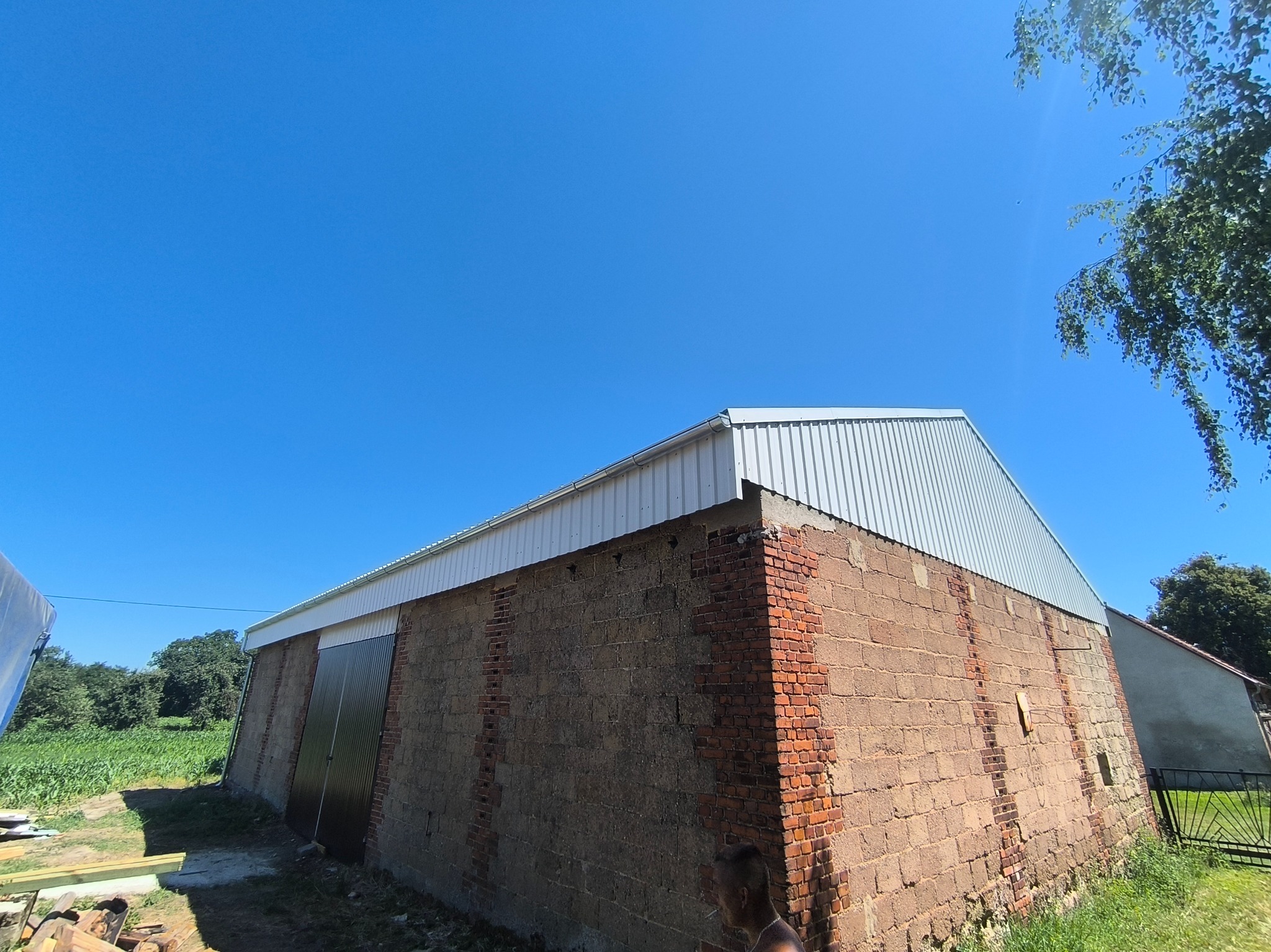 Budynek gospodarczy z nowym, srebrnym dachem trapezowym i ceglanymi ścianami pod błękitnym niebem. Widoczne rynny i fragment zieleni.