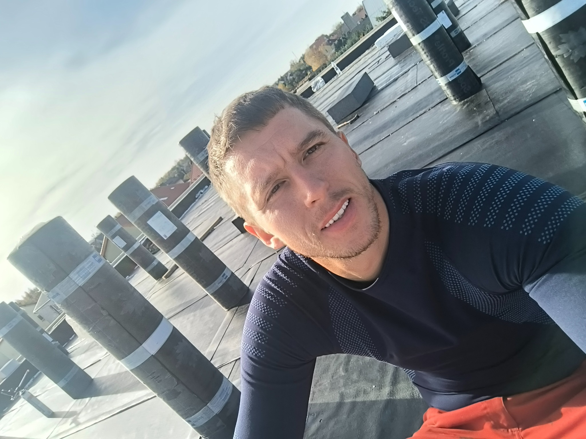 Mężczyzna na dachu pokrytym papą termozgrzewalną, widoczne rolki papy. Ujęcie z dołu, w tle zabudowa miejska i błękitne niebo.