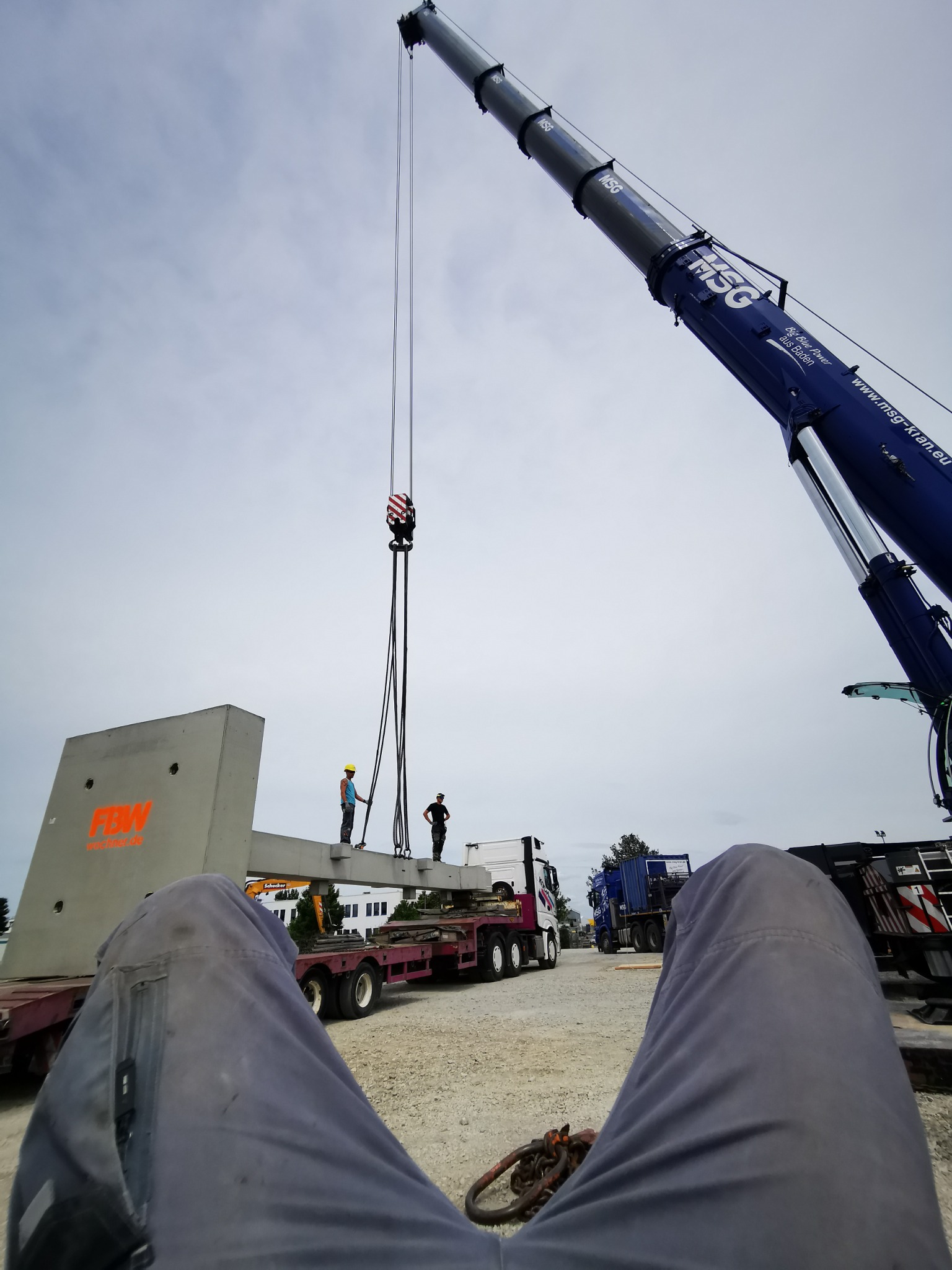 Widok z perspektywy leżącego człowieka na montaż betonowego elementu konstrukcyjnego za pomocą dźwigu, dwóch pracowników w kaskach nadzoruje proces.