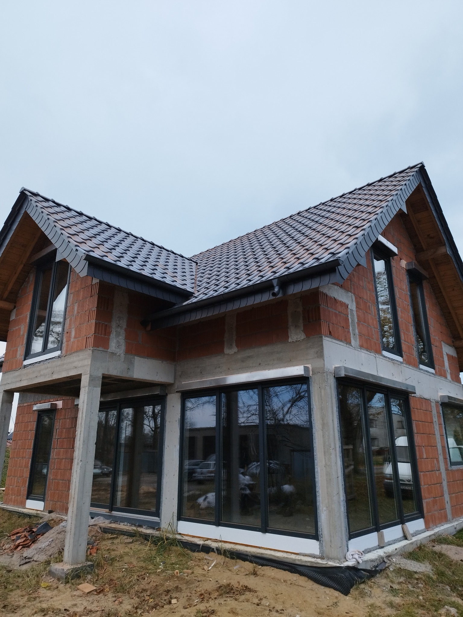 Surowy stan budynku jednorodzinnego z czerwoną cegłą, nowym dachem z ciemnej dachówki ceramicznej i dużymi, czarnymi oknami, w trakcie budowy.
