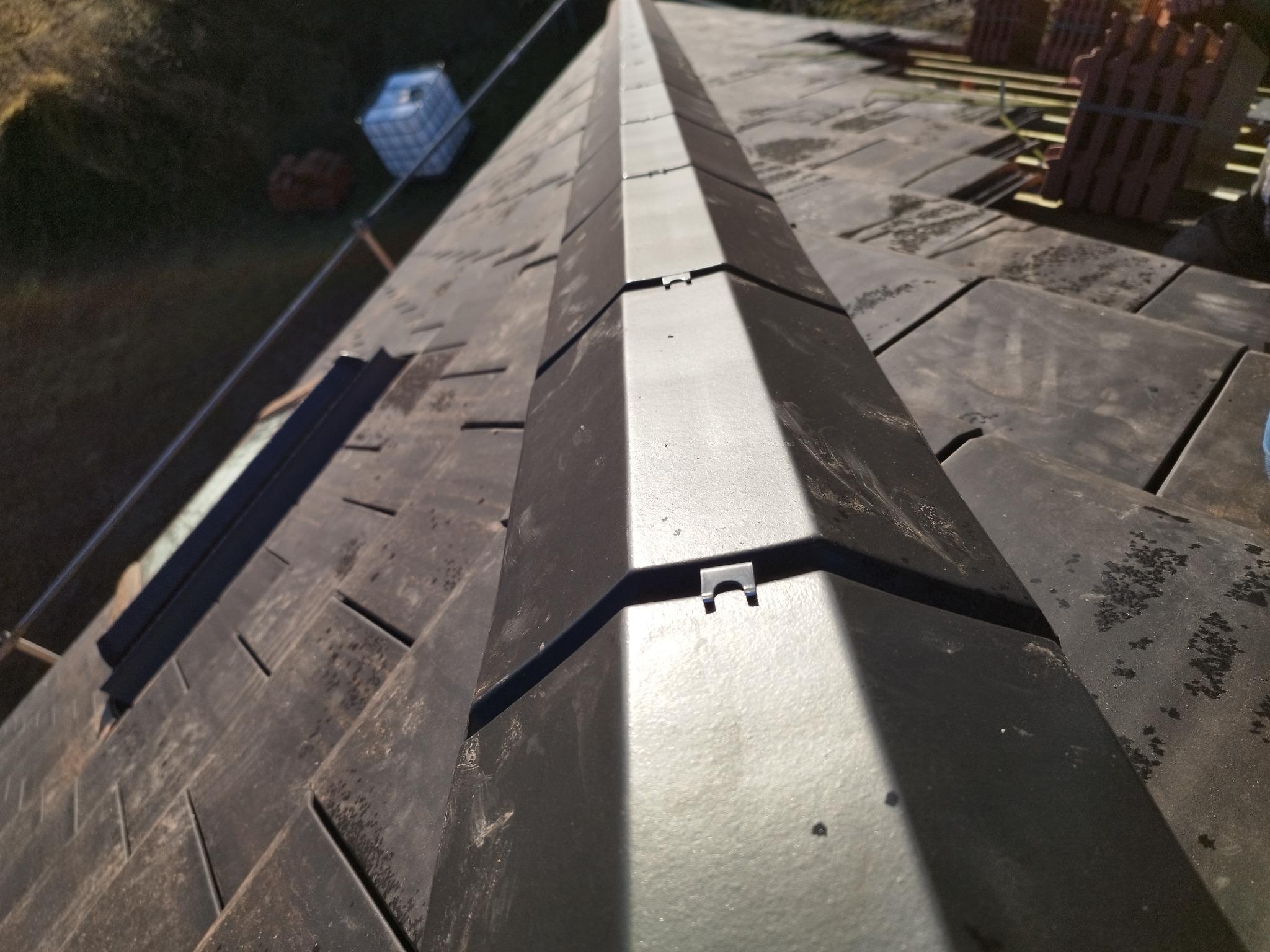 Szary dach z dachówki ceramicznej z widoczną kalenicą, na której widać metalowe uchwyty mocujące, w tle paleta z dachówkami.