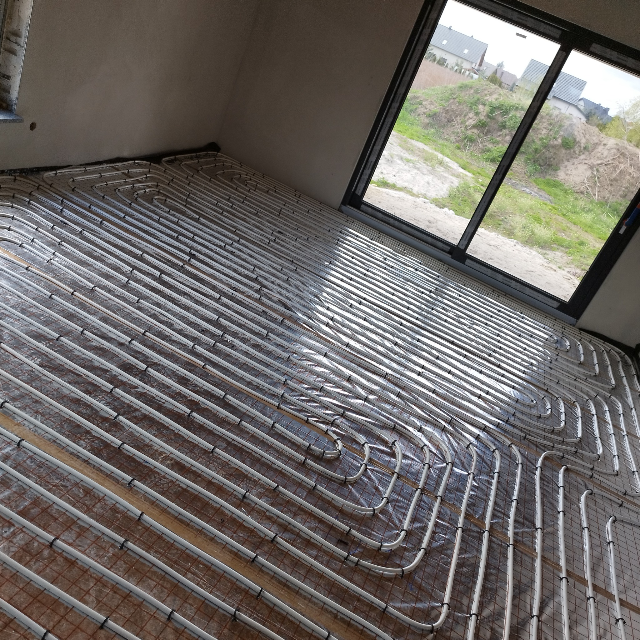 Instalacja ogrzewania podłogowego z widocznymi rurami grzewczymi ułożonymi na siatce, okno w tle z widokiem na zewnątrz.