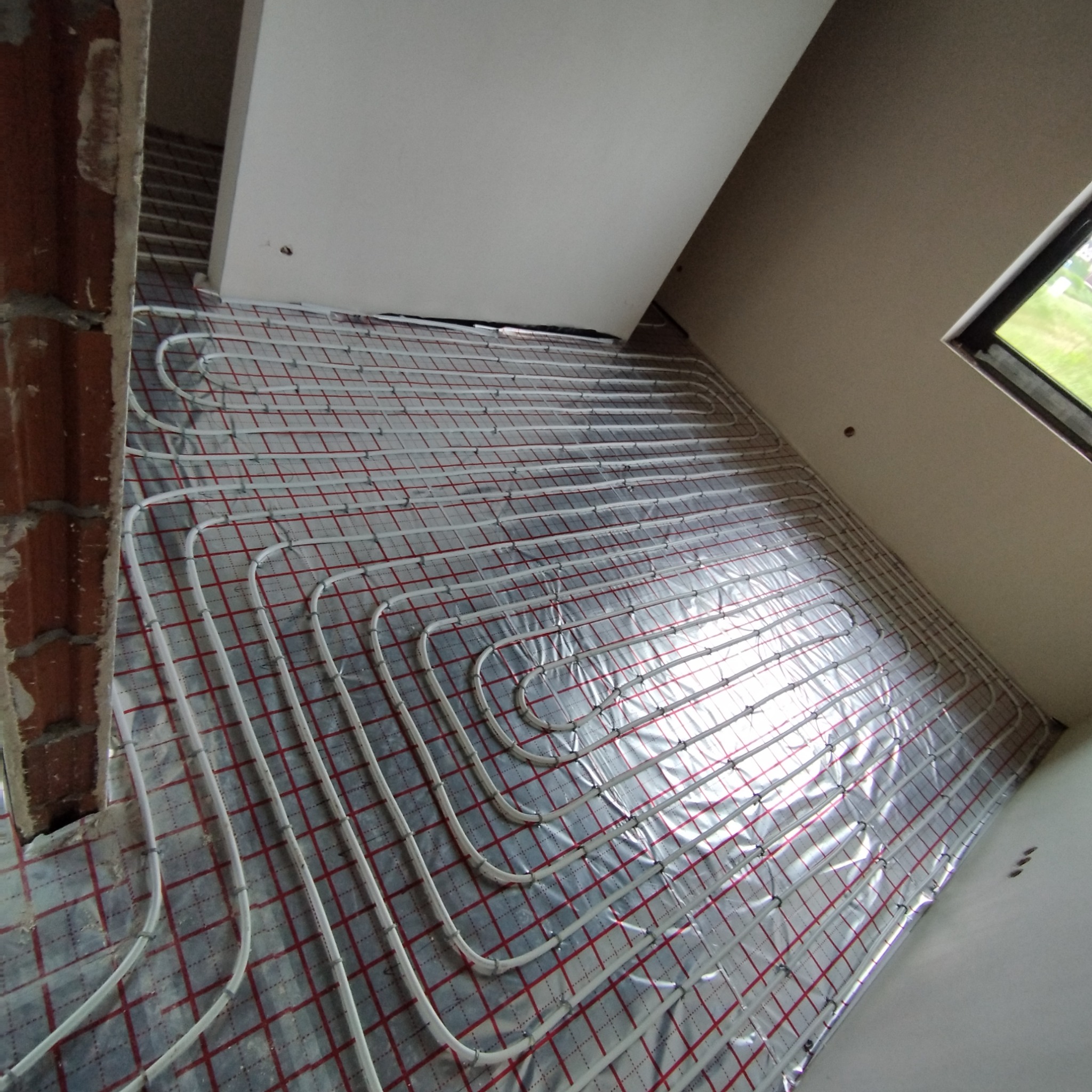 Instalacja ogrzewania podłogowego na folii aluminiowej z czerwoną siatką, widoczne rury w kształcie meandrów, ściany w stanie surowym i okno z widokiem na zieleń.