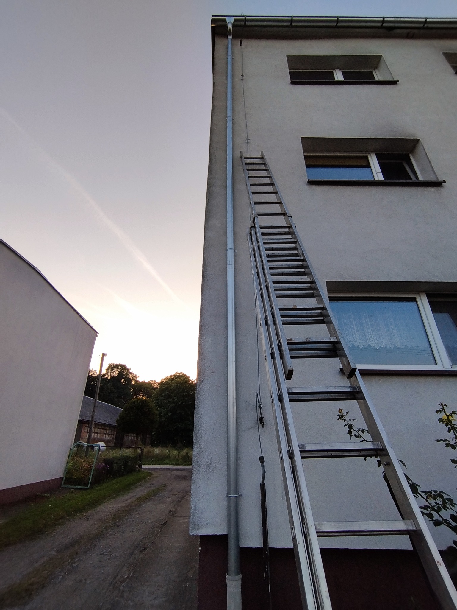 Metalowa drabina oparta o ścianę budynku z rynną, widoczna droga gruntowa i zieleń w tle, wieczorny krajobraz.