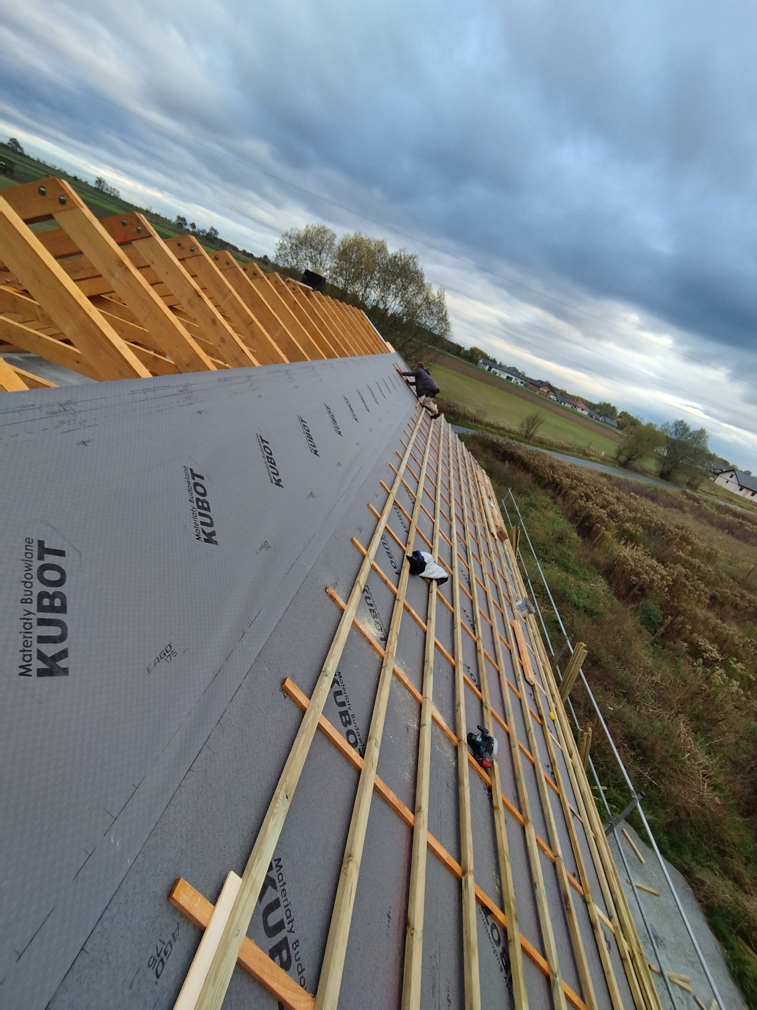 Konstrukcja drewniana dachu pokryta membraną KUBOT, widok z góry na rozległe pola i zabudowania w oddali, prace dekarskie w toku.
