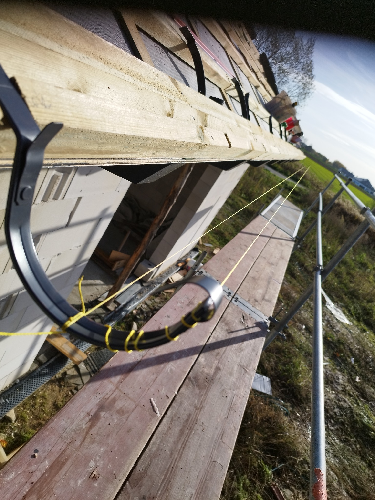 Montaż rynny dachowej z użyciem drabiny i sznurka poziomującego, widoczny fragment dachu z deskowaniem i membraną, oraz fragment elewacji budynku.