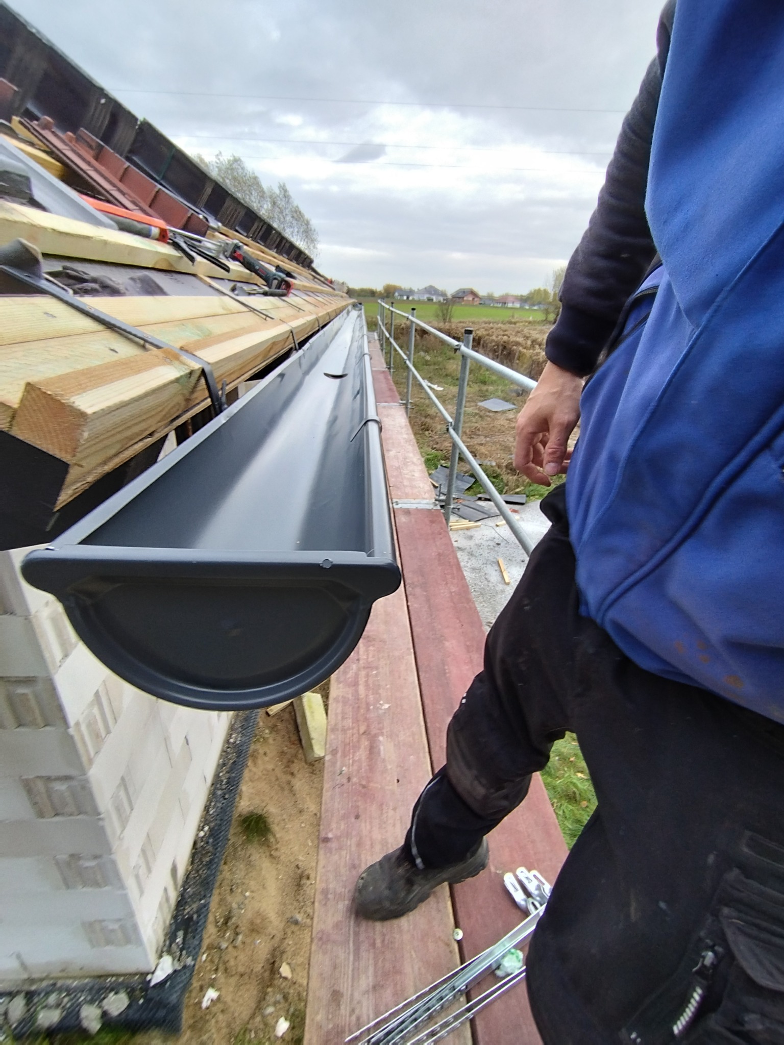 Montaż szarej rynny dachowej, widok z góry na krawędź dachu z drewnianymi elementami konstrukcyjnymi, biały mur i rusztowanie w tle.